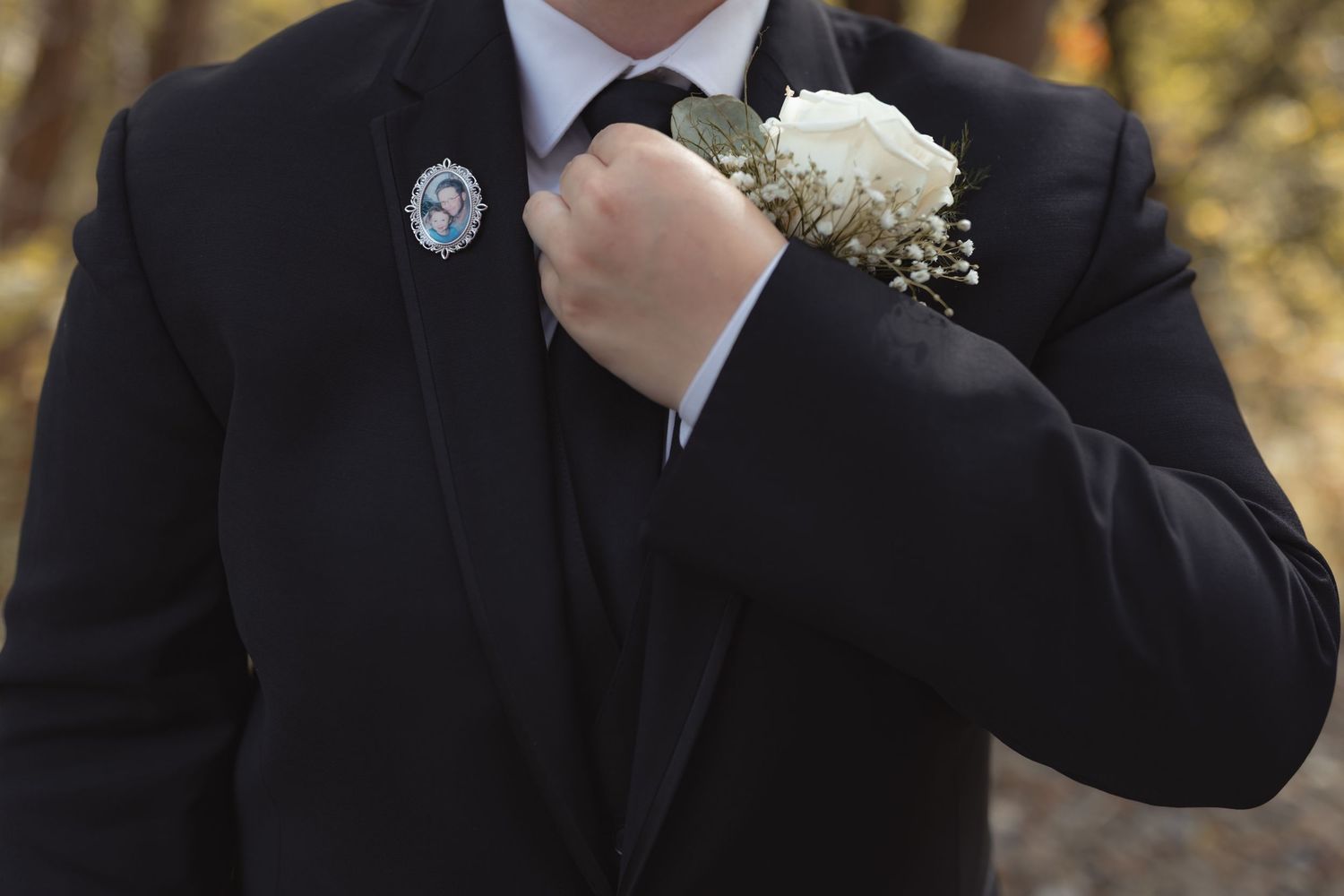 Groom in black suit adjusting boutonniere with memorial pin on lapel.