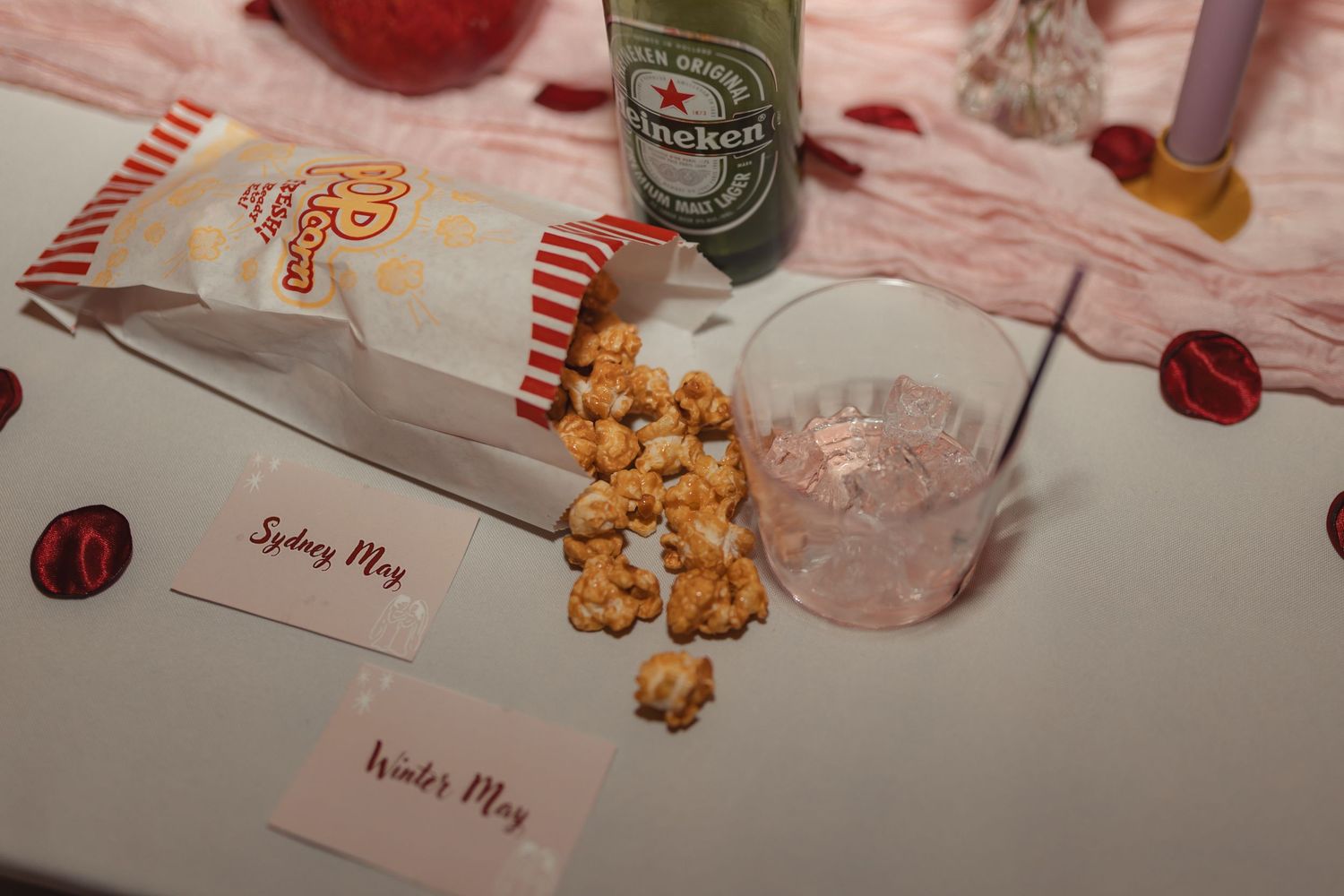 Caramel popcorn spilled from a striped bag beside a pink drink, Heineken bottle, and name cards.
