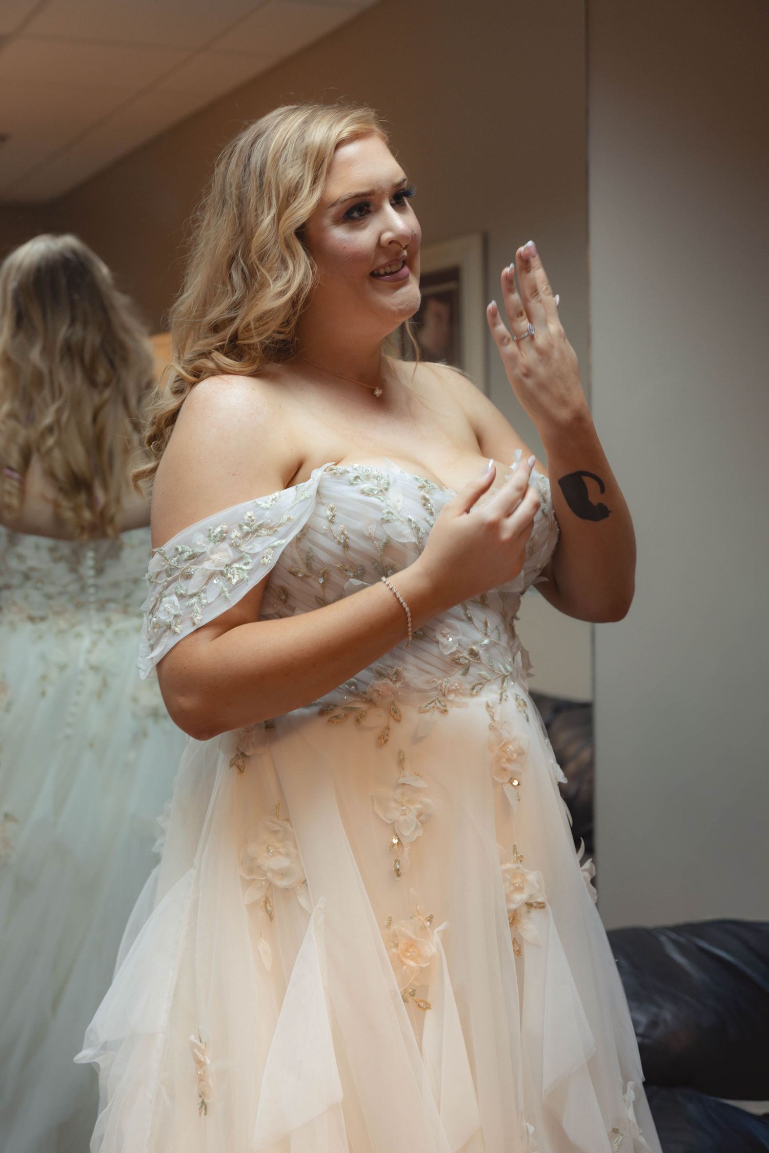 Blonde bride in off-shoulder floral tulle wedding gown admiring her ring in bridal suite mirror.