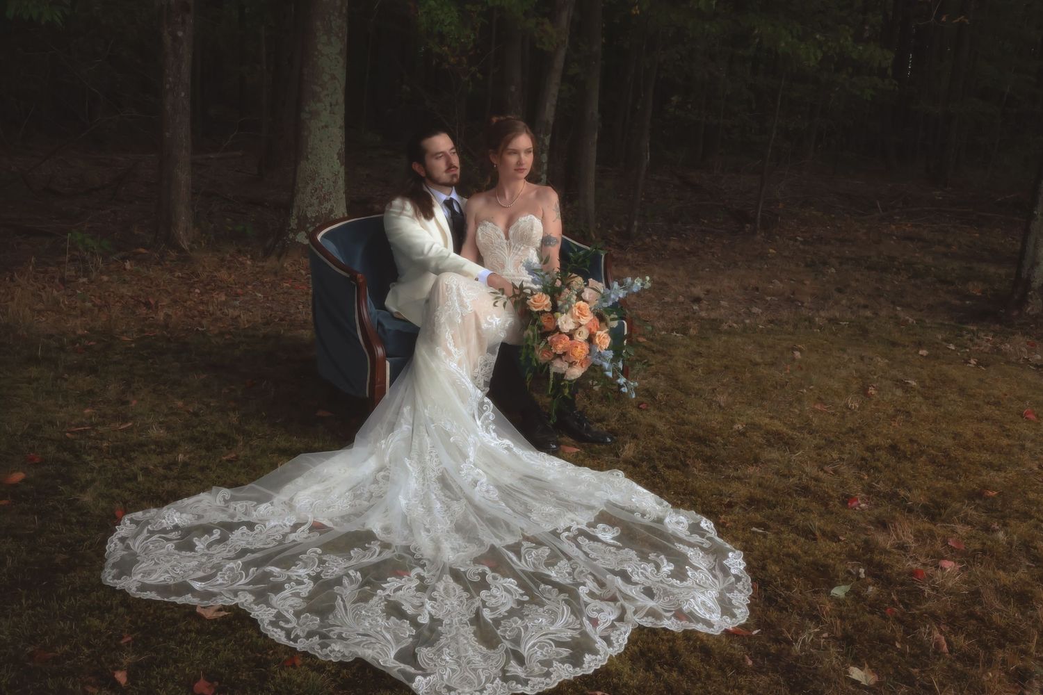 Bride and groom pose in forest, her lace gown train spread wide, holding autumn floral bouquet.