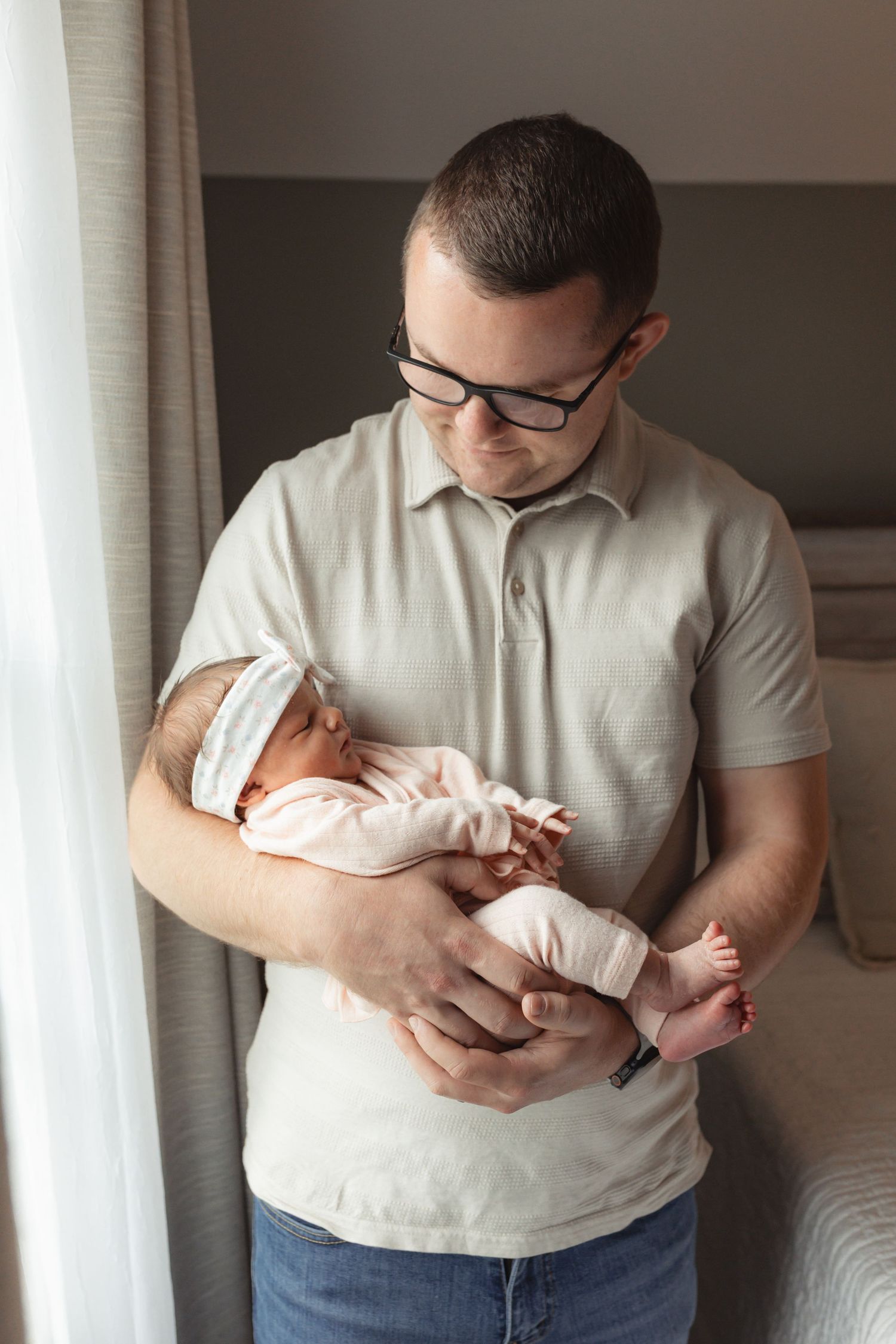 A new father wearing glasses tenderly holds his sleeping newborn baby in pink clothing near a window.