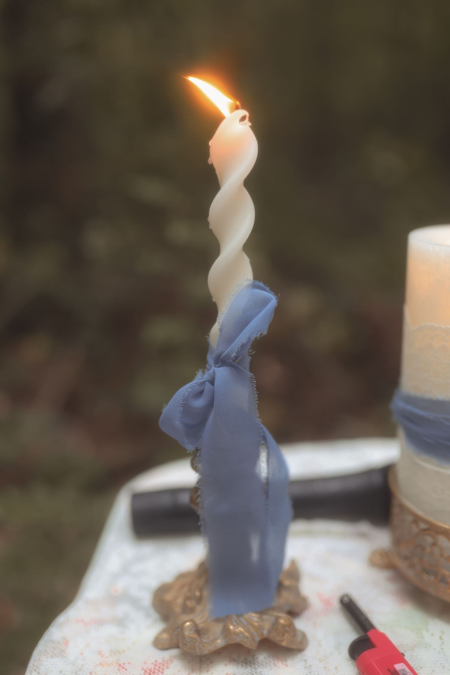 A twisted white and blue decorative candle with a gold base burning outdoors at a wedding ceremony.