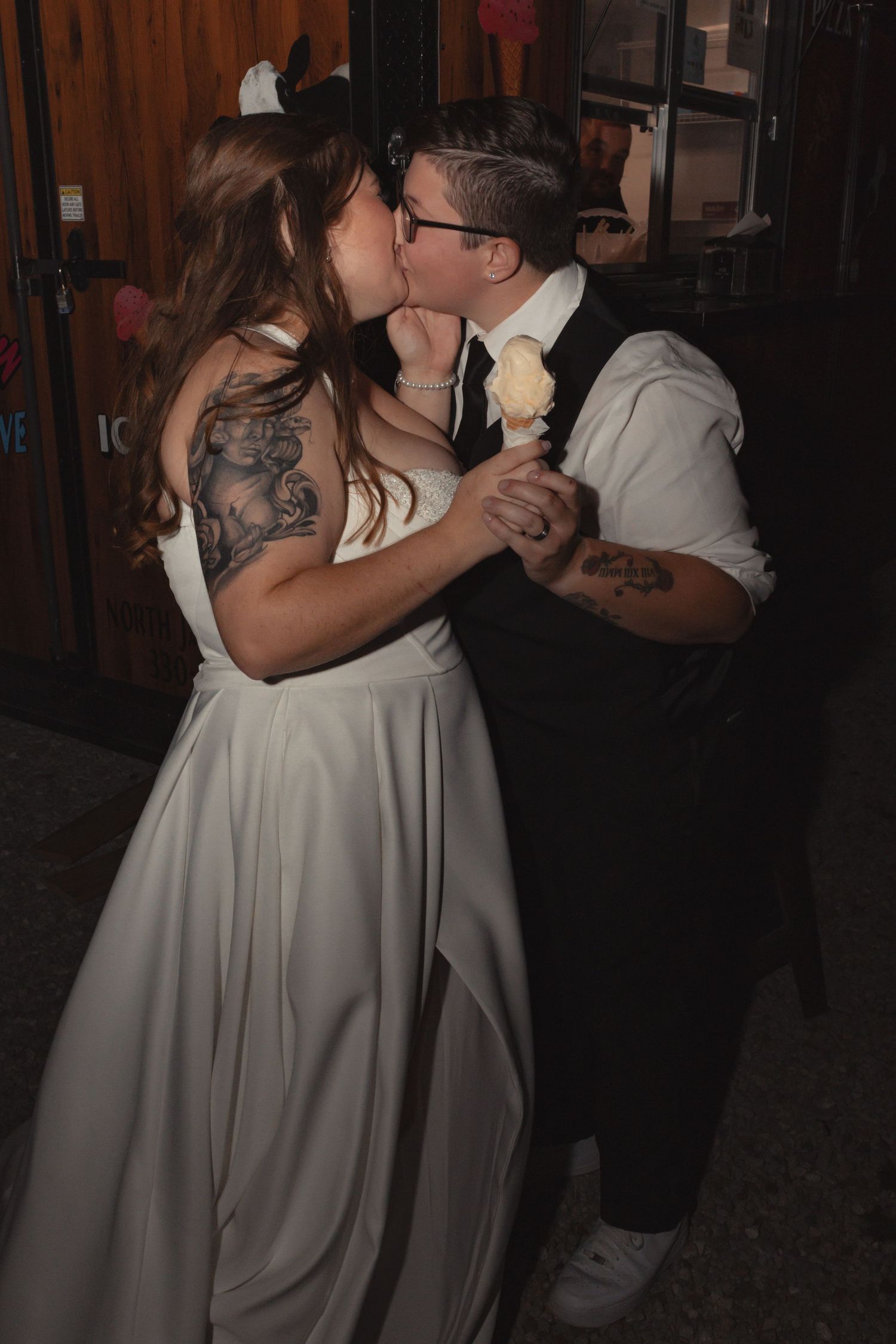 A bride and groom share a kiss while dancing, bride in white gown with tattoos, groom in vest holding a flower.