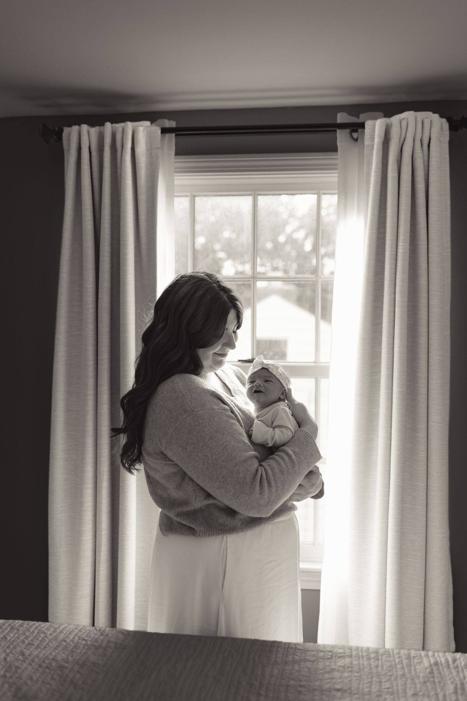 Black and white photo of a mother tenderly holding her newborn baby by a bright window with white curtains.