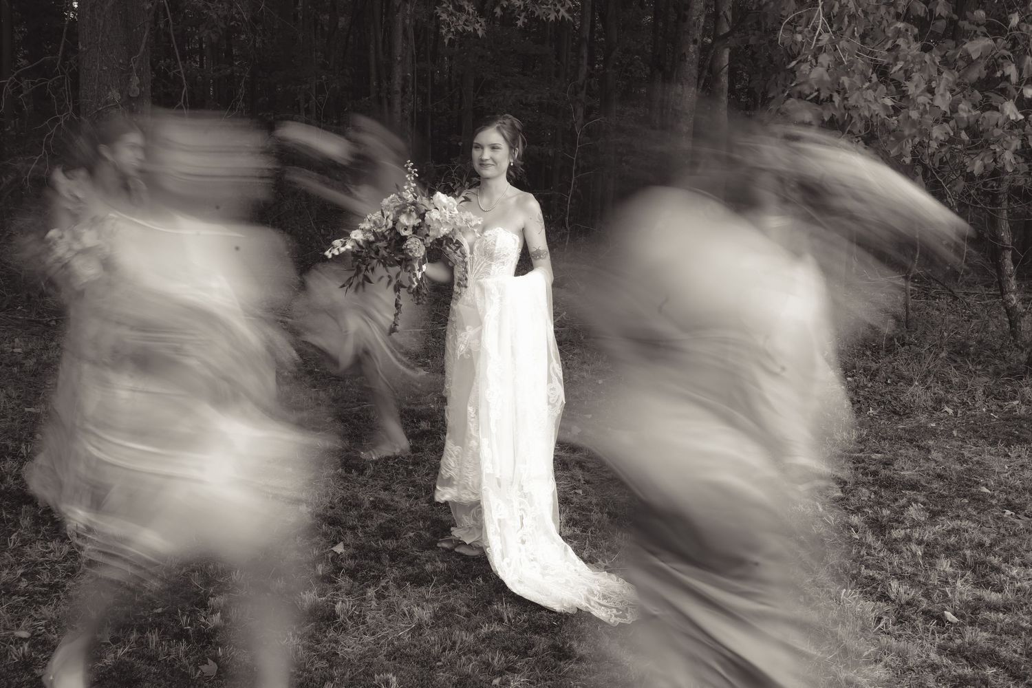 Bride in white gown holds bouquet in forest while blurred wedding party moves around her in black and white photo.