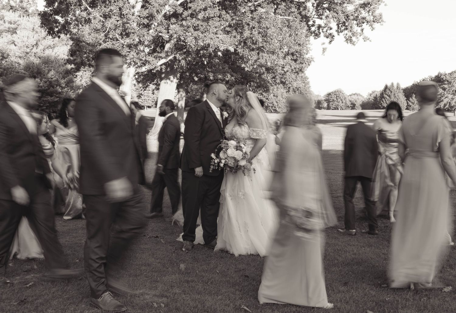 Black and white wedding photo of bride and groom surrounded by guests outdoors under a tree.