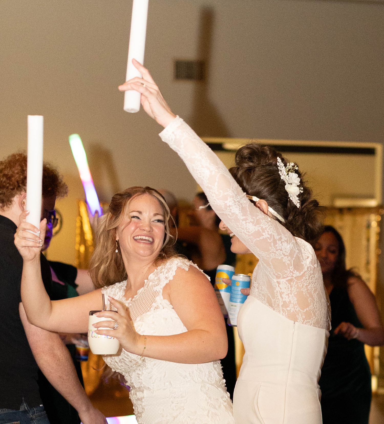 Two brides in white lace gowns celebrate at their wedding reception, dancing with glow sticks.
