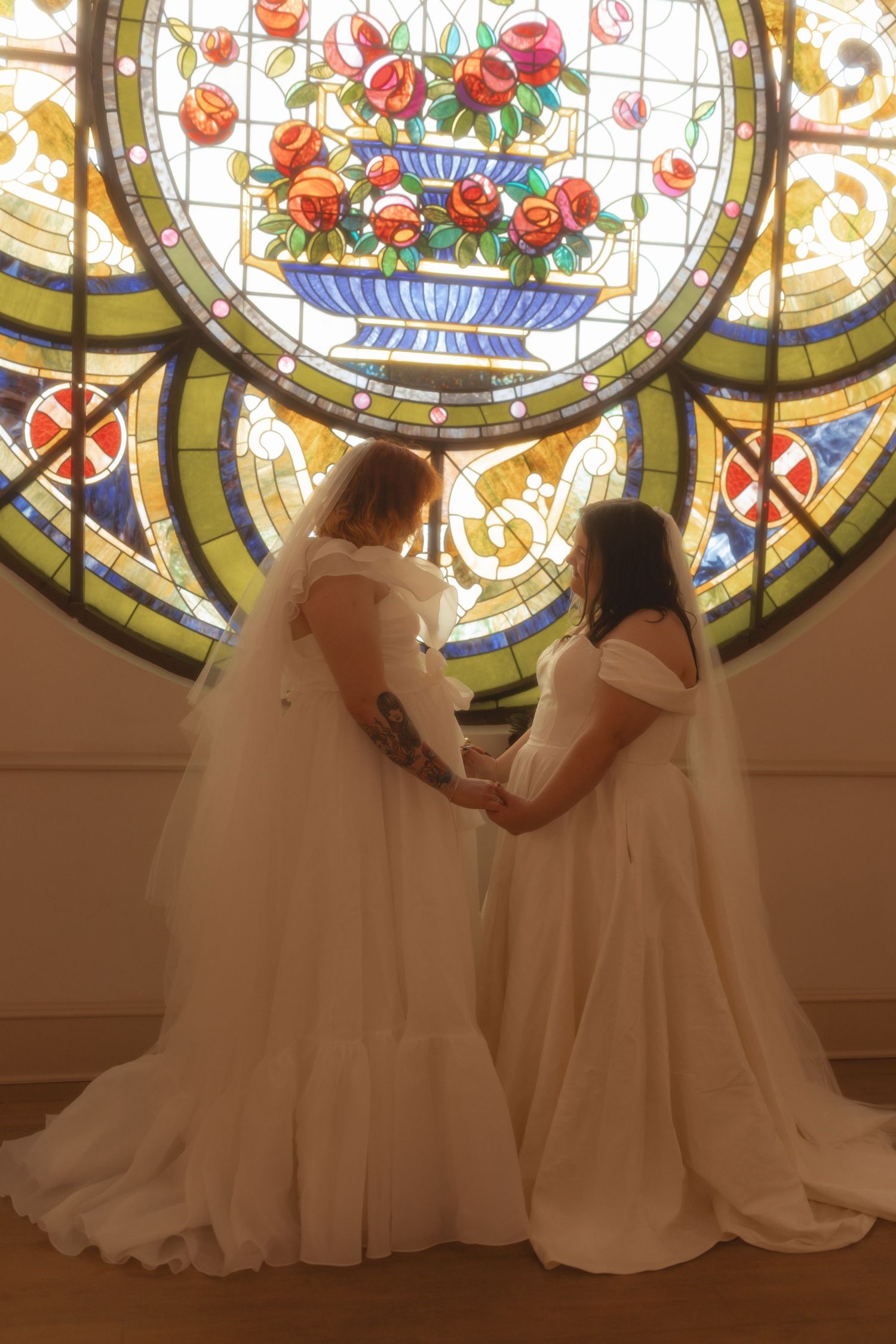 Two brides in white gowns hold hands before a stunning circular stained glass window featuring roses.