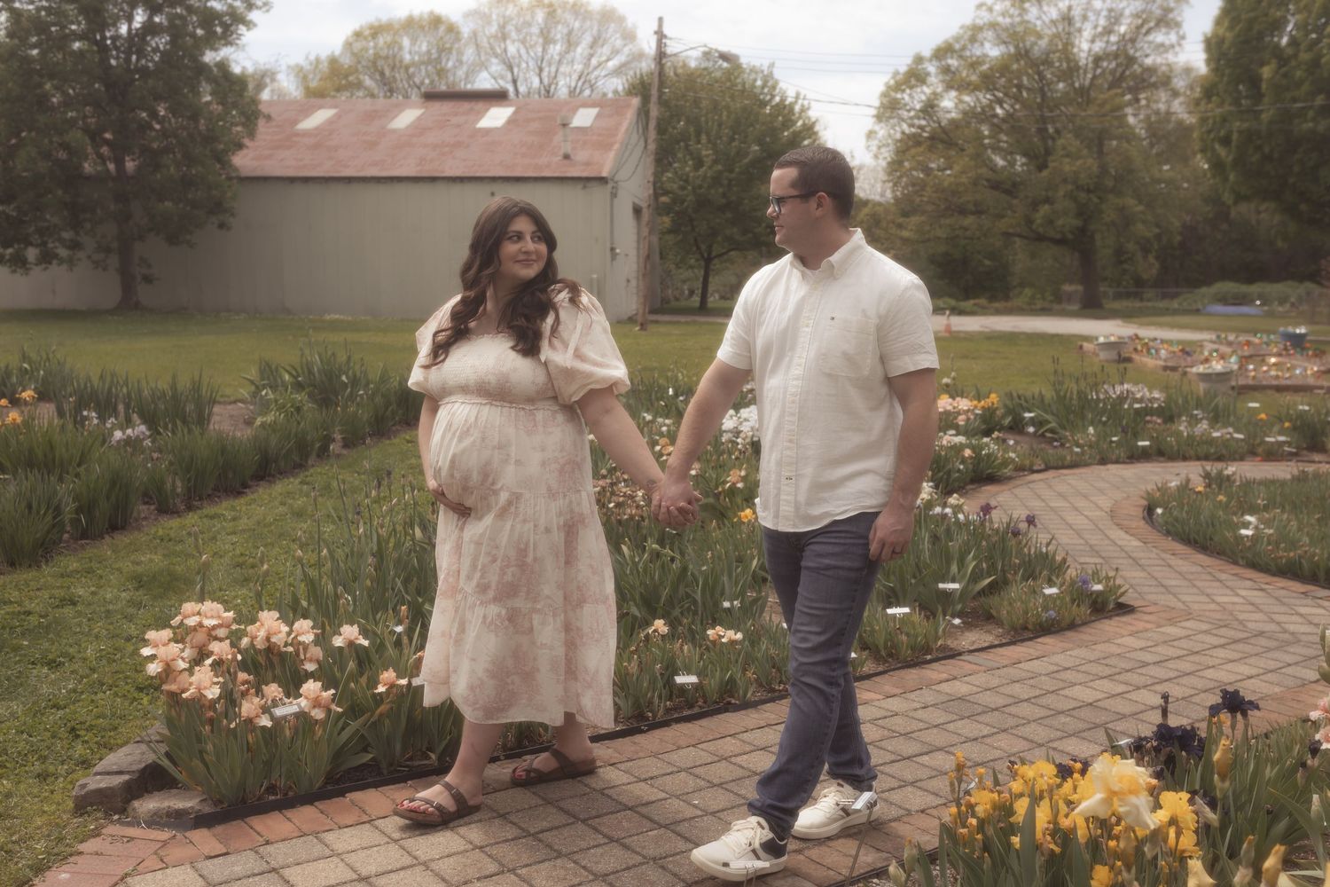 Pregnant woman in floral dress holding hands with man walking through colorful garden path.