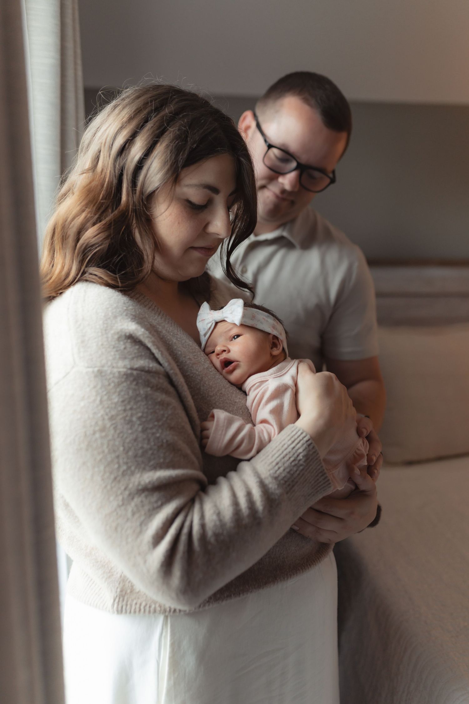 Parents lovingly hold their swaddled newborn baby girl with a white bow headband during an in-home session.
