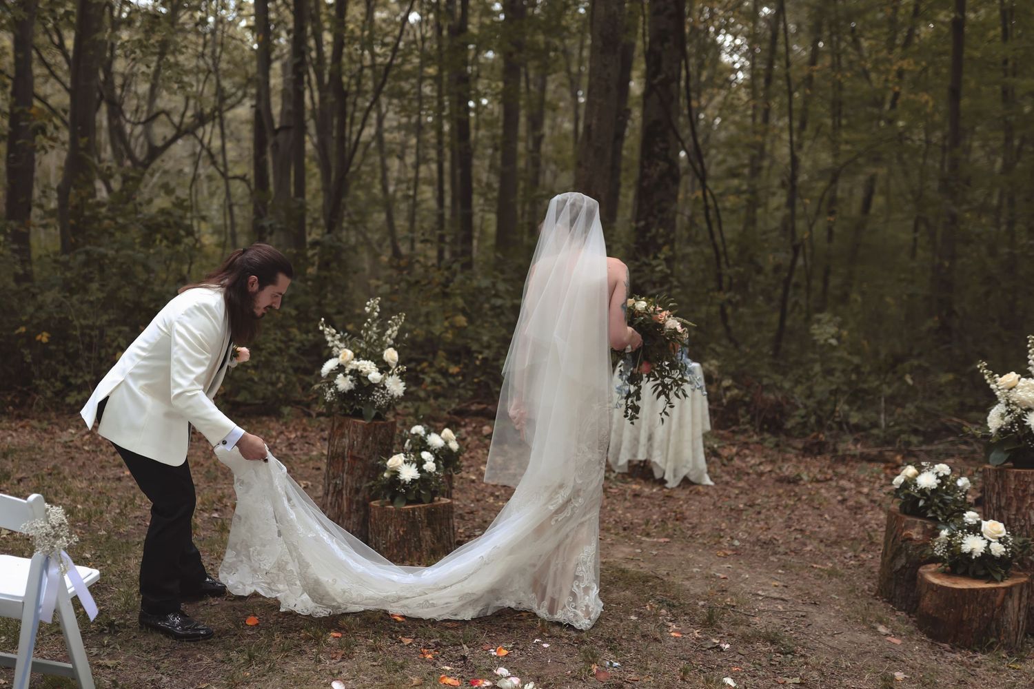 Groom in white jacket arranging bride's lace wedding veil in a romantic woodland forest ceremony setting.