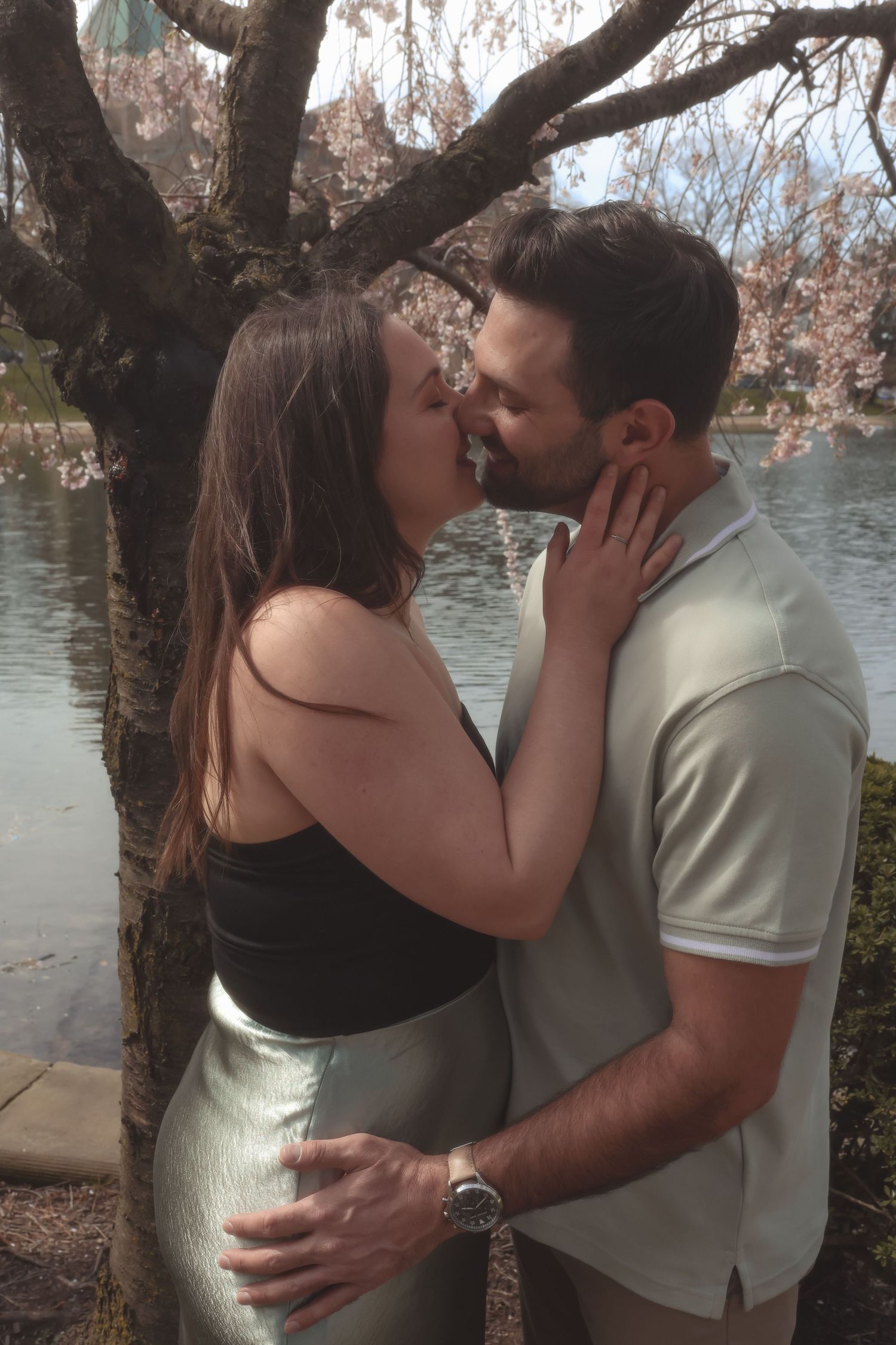 Couple sharing a romantic kiss under blooming cherry blossom tree by a serene lake in spring.