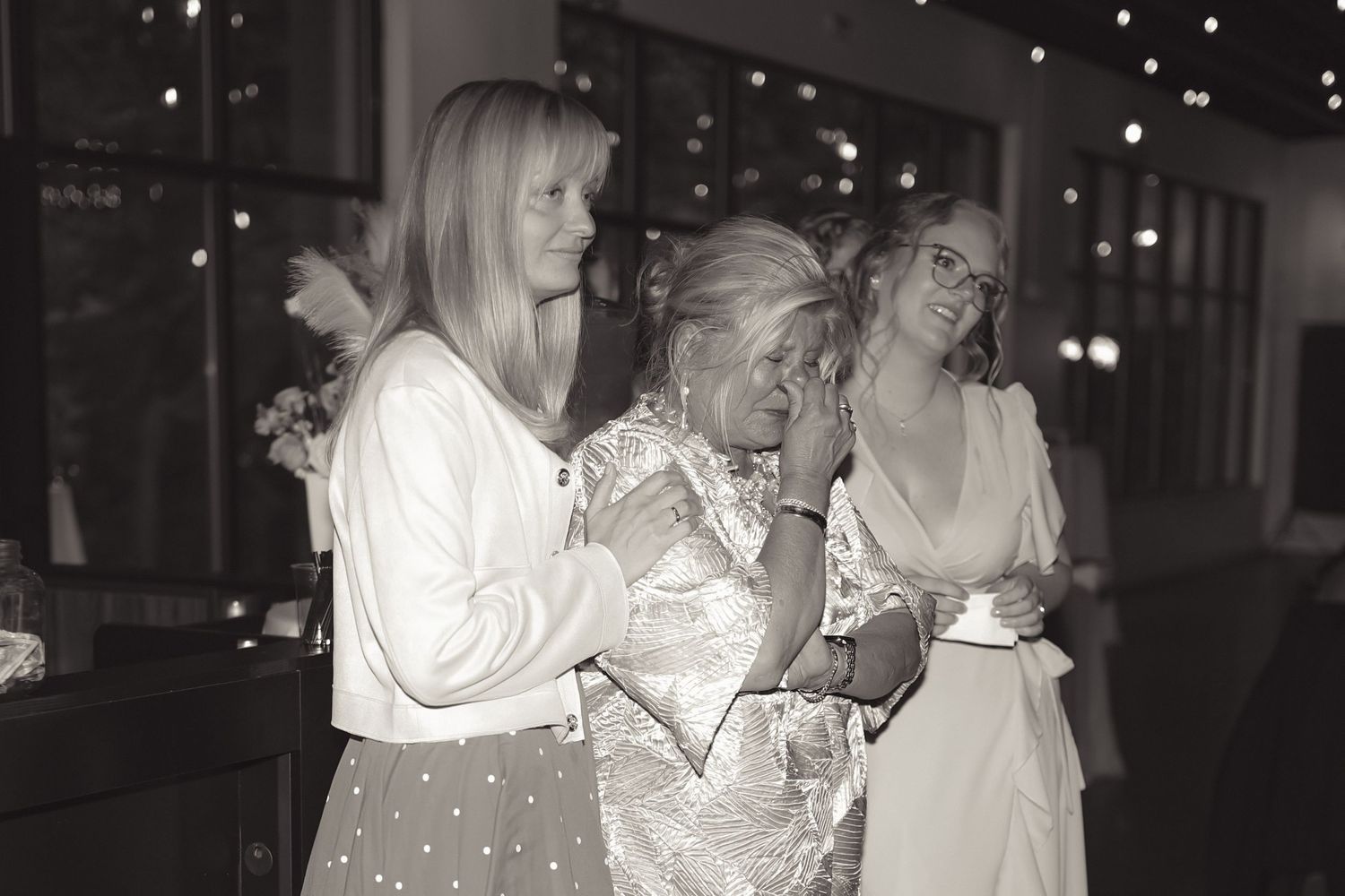Black and white photo of three women at a wedding reception, with the middle woman wiping tears of joy.