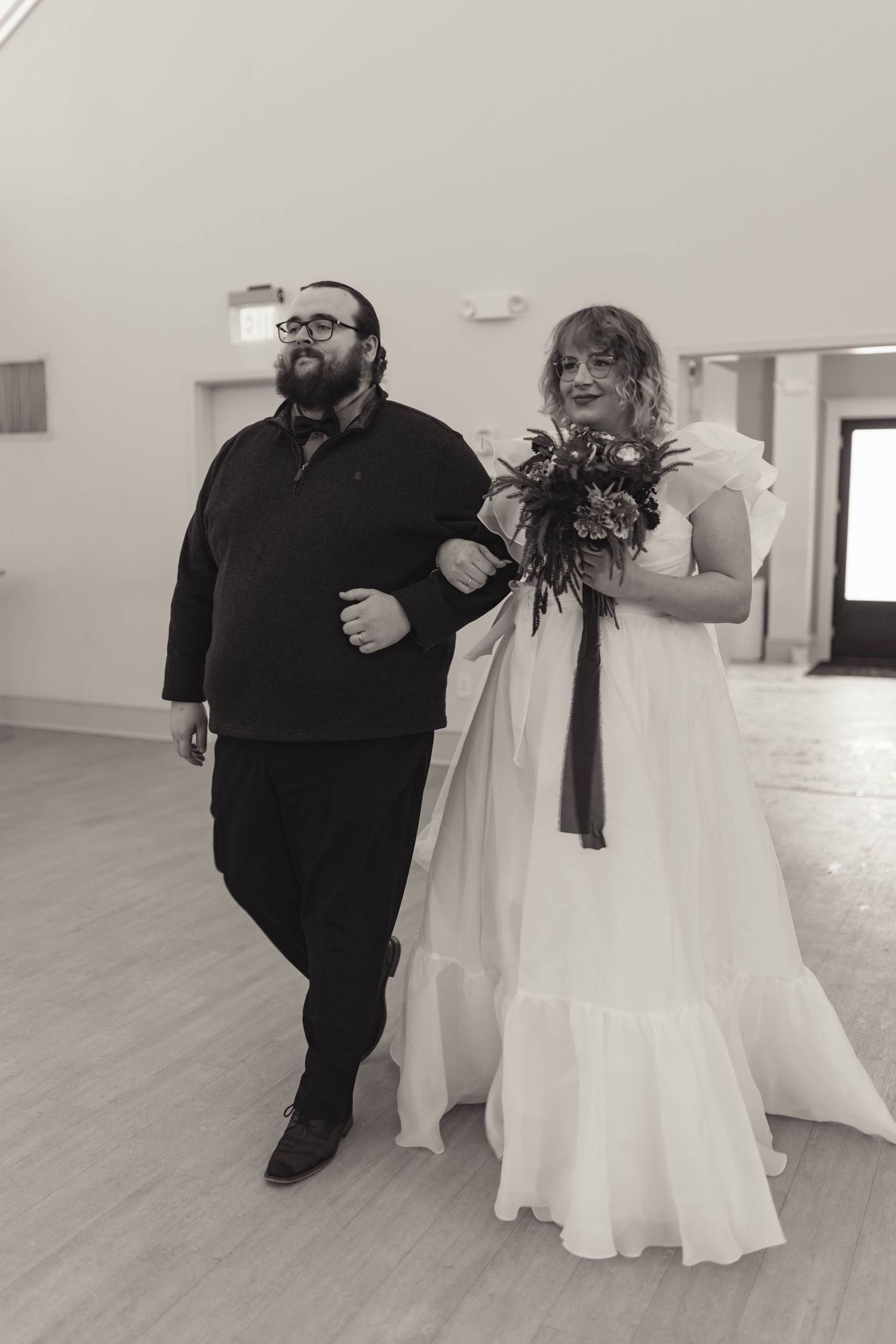 Black and white photo of a bride in ruffled gown with bouquet walking arm-in-arm with a man.