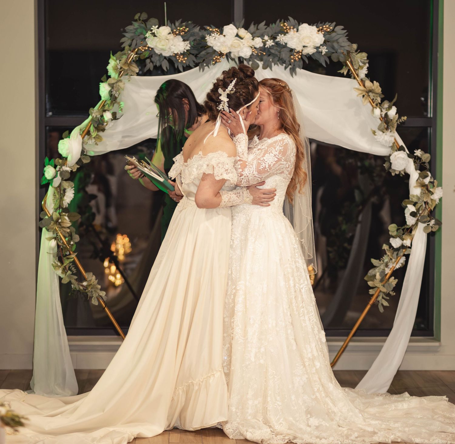 Two brides sharing a kiss under a gold arch adorned with white and green flowers during their wedding ceremony.