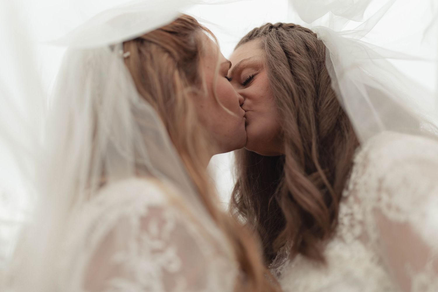 Two brides sharing a romantic kiss under wedding veils in a soft, ethereal bridal portrait.