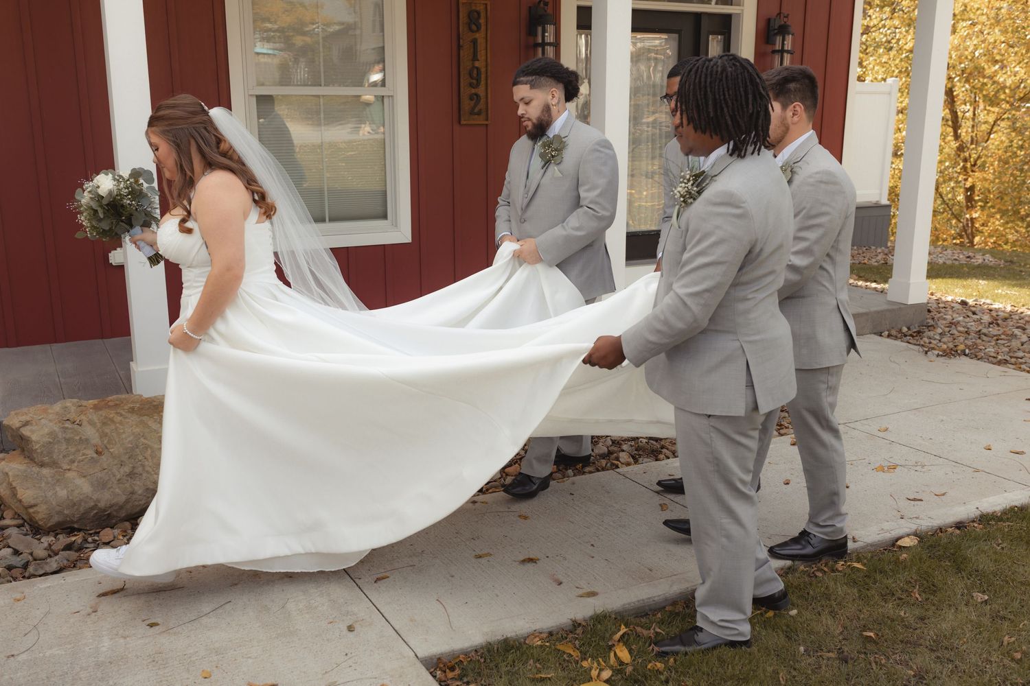 Groomsmen in grey suits help carry bride's white wedding dress train outside red barn wedding venue.