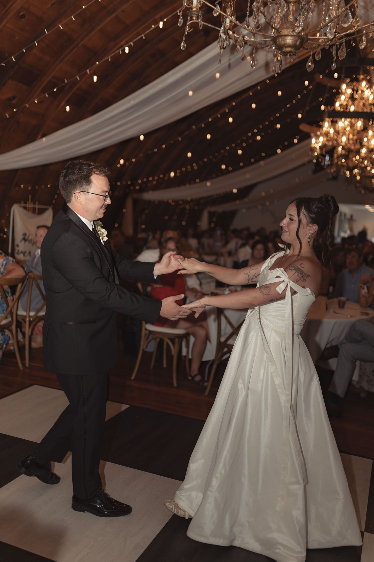 Bride and groom share their first dance in a warmly lit wedding venue decorated with string lights and chandeliers.