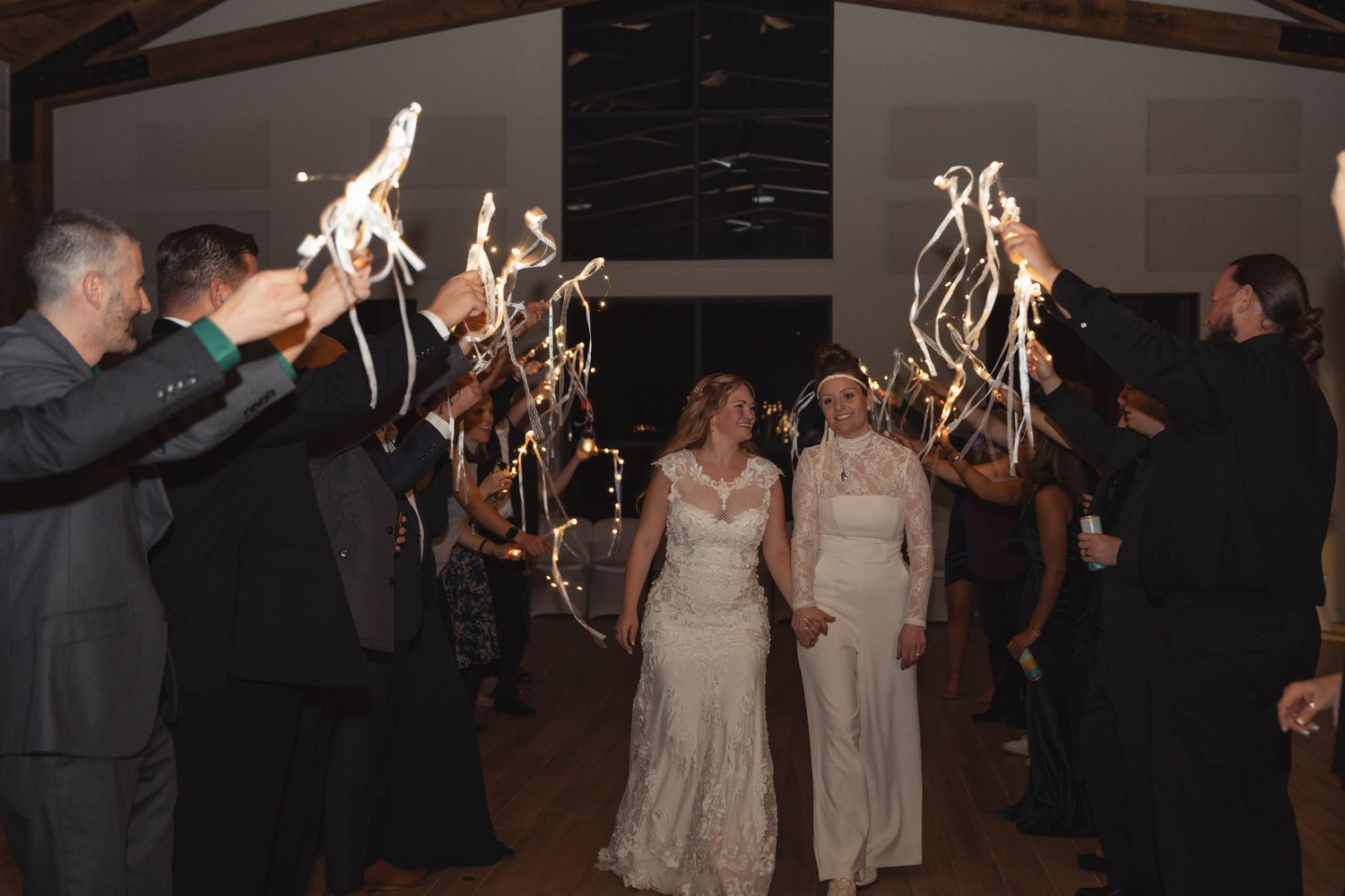 Two brides walk through a sparkler exit tunnel at their wedding reception, surrounded by cheering guests.