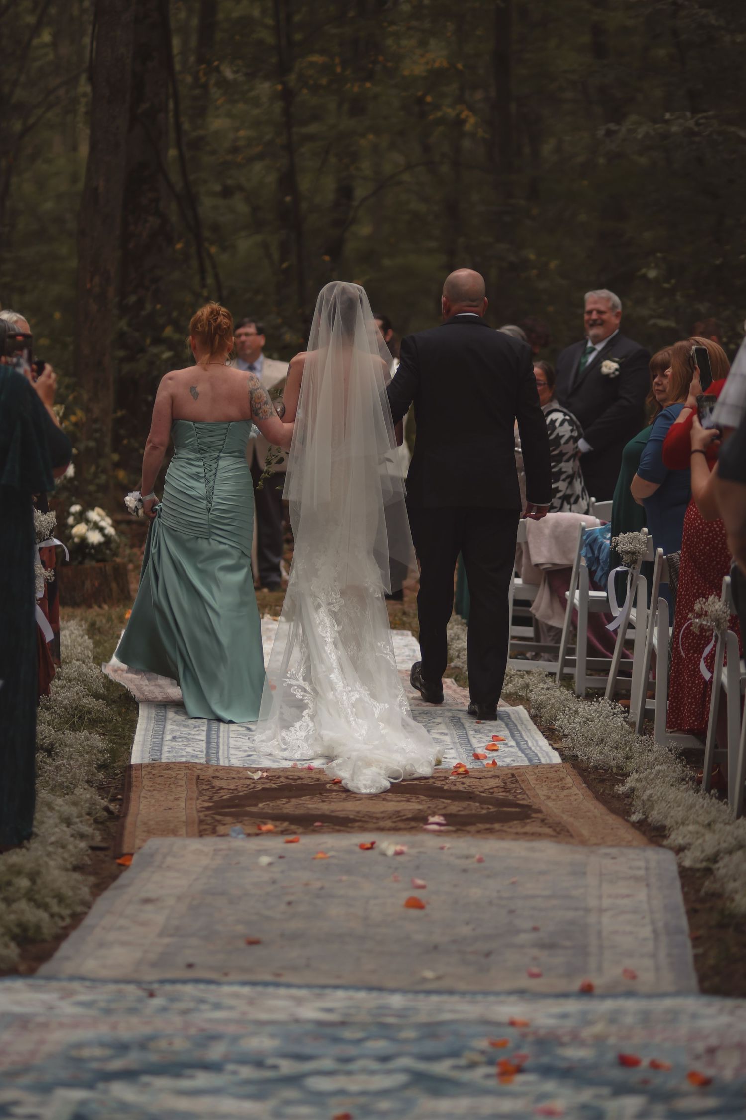 Bride walks down rustic outdoor aisle in forest wedding, escorted by two people past seated guests.