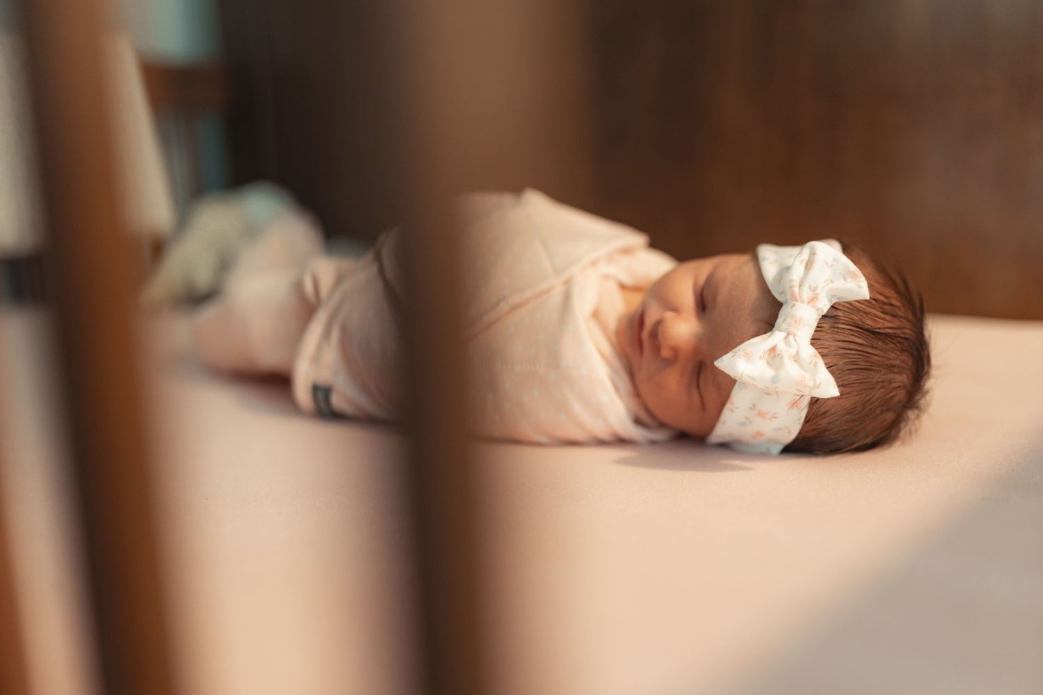 Newborn baby swaddled in pink sleeping peacefully in a crib wearing a white bow headband.