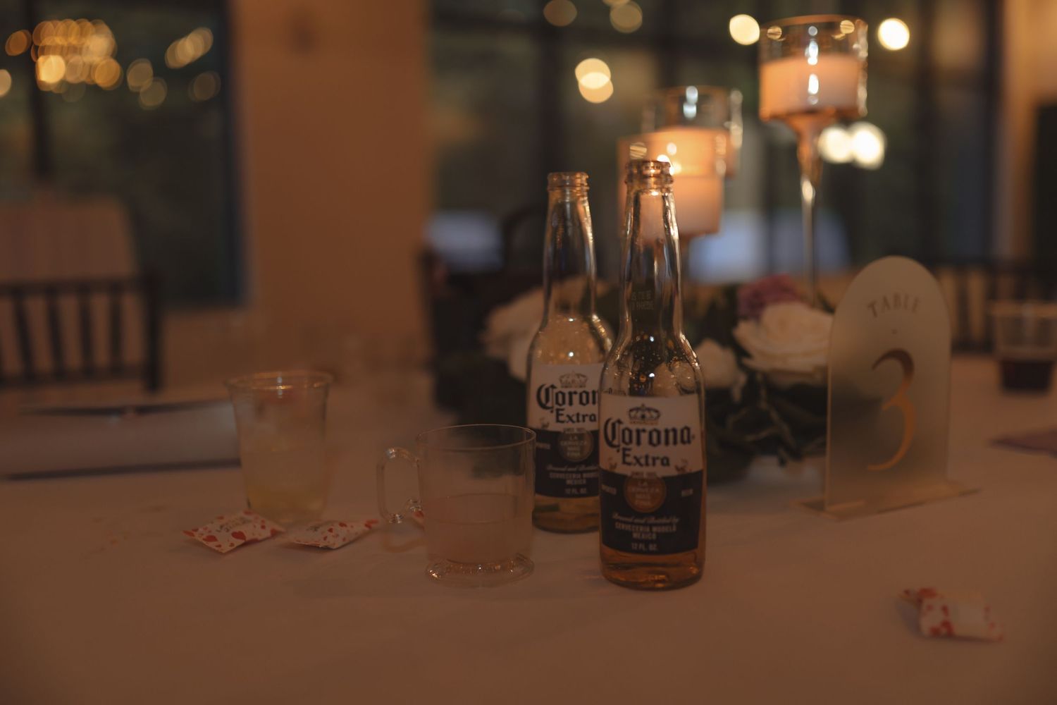 Two Corona Extra beer bottles on a dimly lit wedding reception table with candles and glassware.