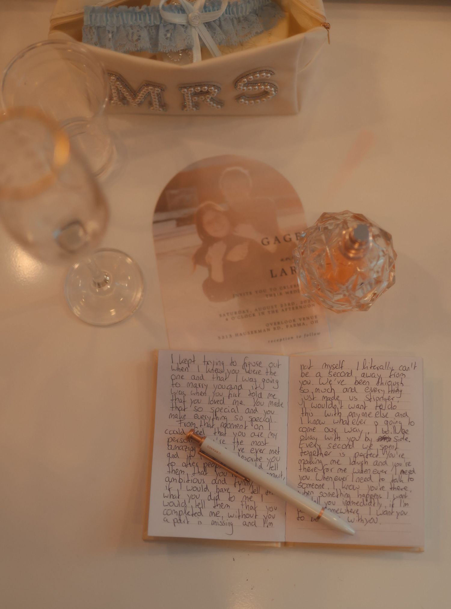 A wedding flat lay featuring a handwritten letter, heart-shaped invitation, MRS tiara, perfume bottle, and wine glass on white surface.