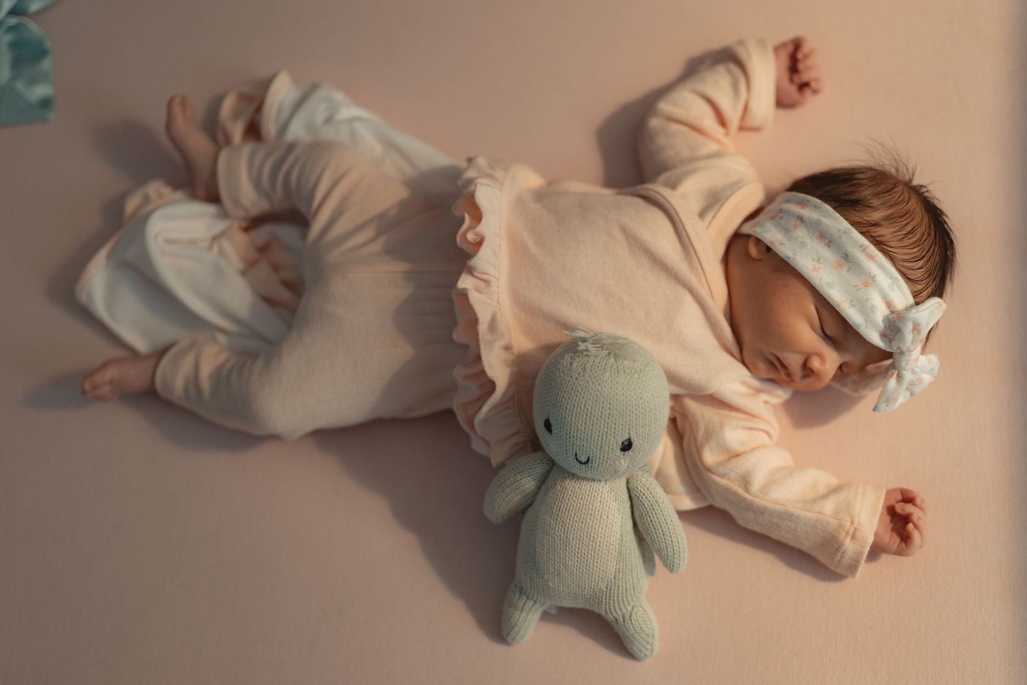 Newborn baby sleeping peacefully in pink outfit with a soft blue bunny stuffed animal toy on a pink background.