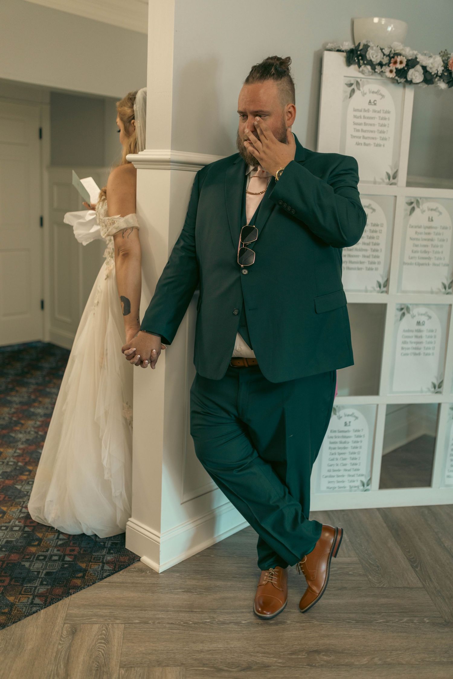 Groom in teal suit holds bride's hand around a white pillar, covering his emotional reaction on their wedding day.