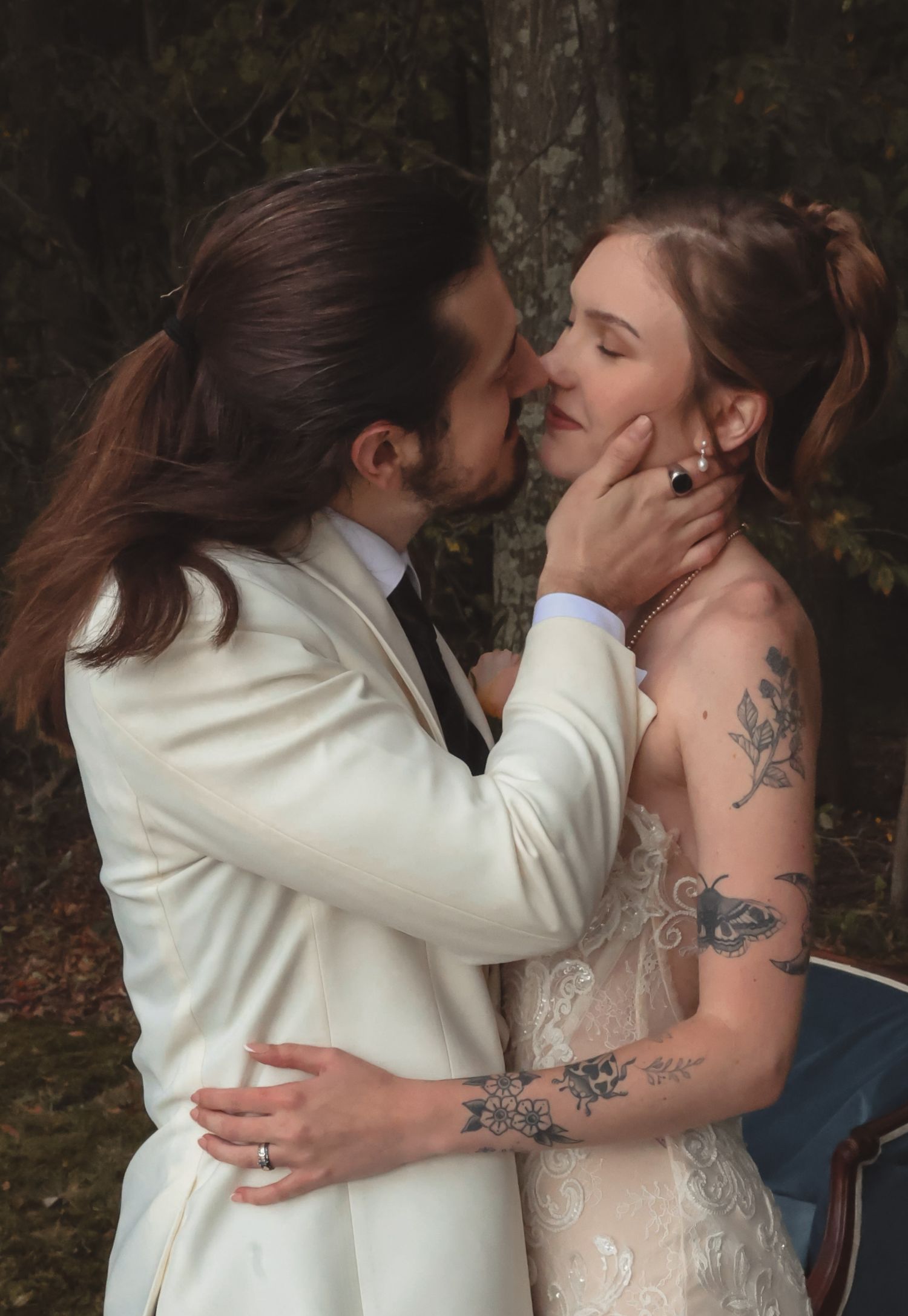 A couple shares a romantic kiss in wedding attire, the bride with tattoos in lace gown, groom in white suit outdoors.