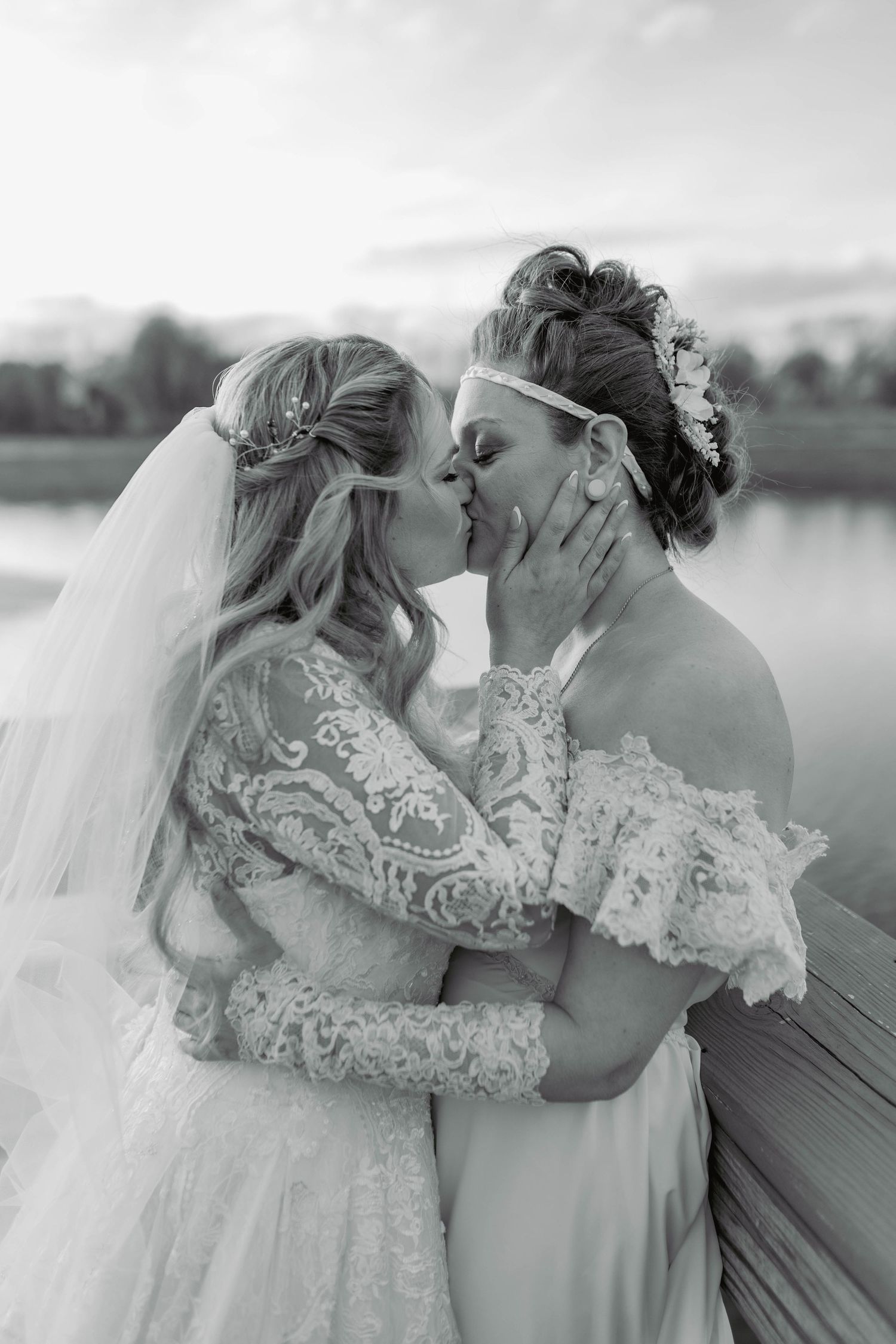 Two brides in white lace wedding dresses share a romantic kiss by a lakeside in black and white.