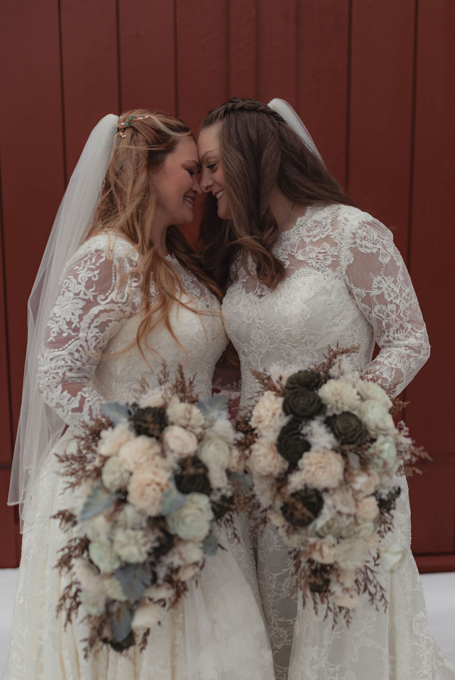 Two brides in lace wedding dresses holding bouquets of white and sage flowers, touching foreheads and smiling.