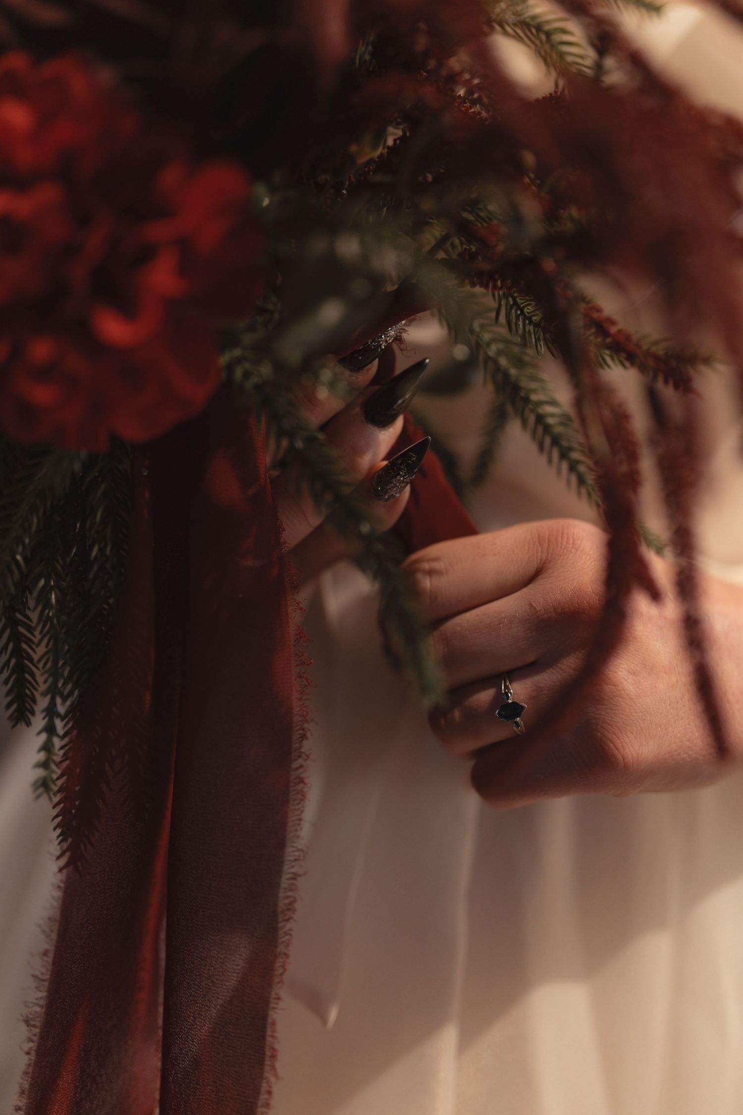 Close-up of a bride holding a dark red wedding bouquet with greenery, wearing a black gemstone ring and dark nails.