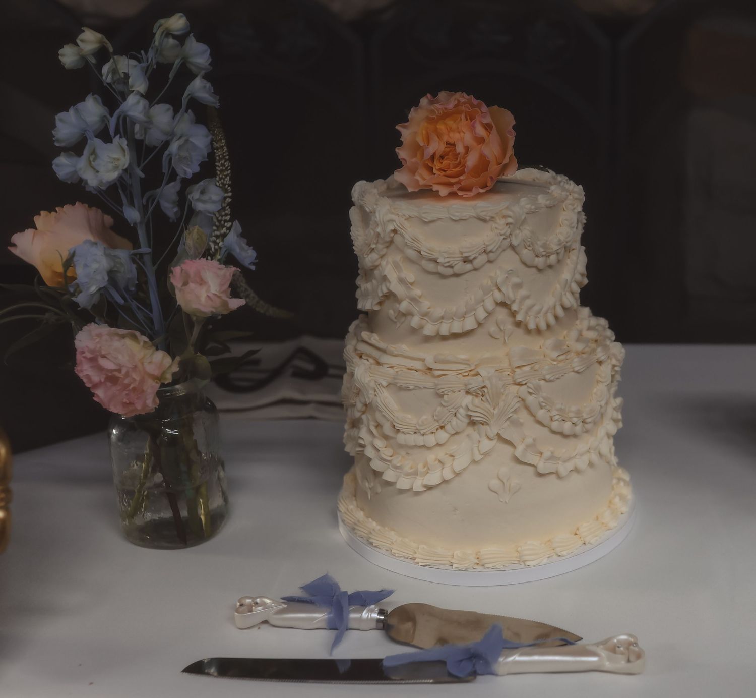 Elegant two-tier white wedding cake with ruffled frosting and peach flower topper beside a floral arrangement.