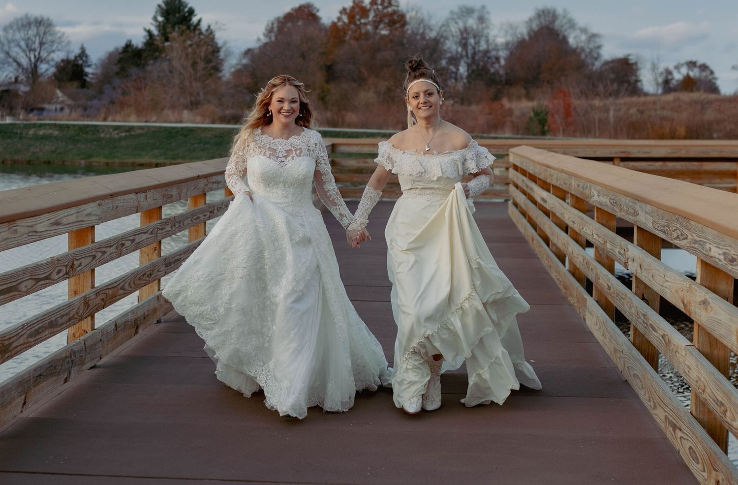 Two brides in white wedding gowns hold hands on a wooden bridge surrounded by autumn trees.