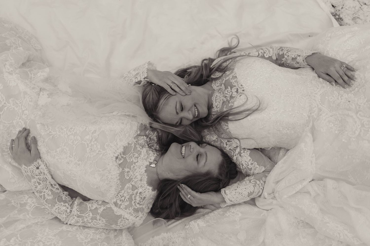 Two brides in lace gowns lie together laughing in a black and white wedding photo.