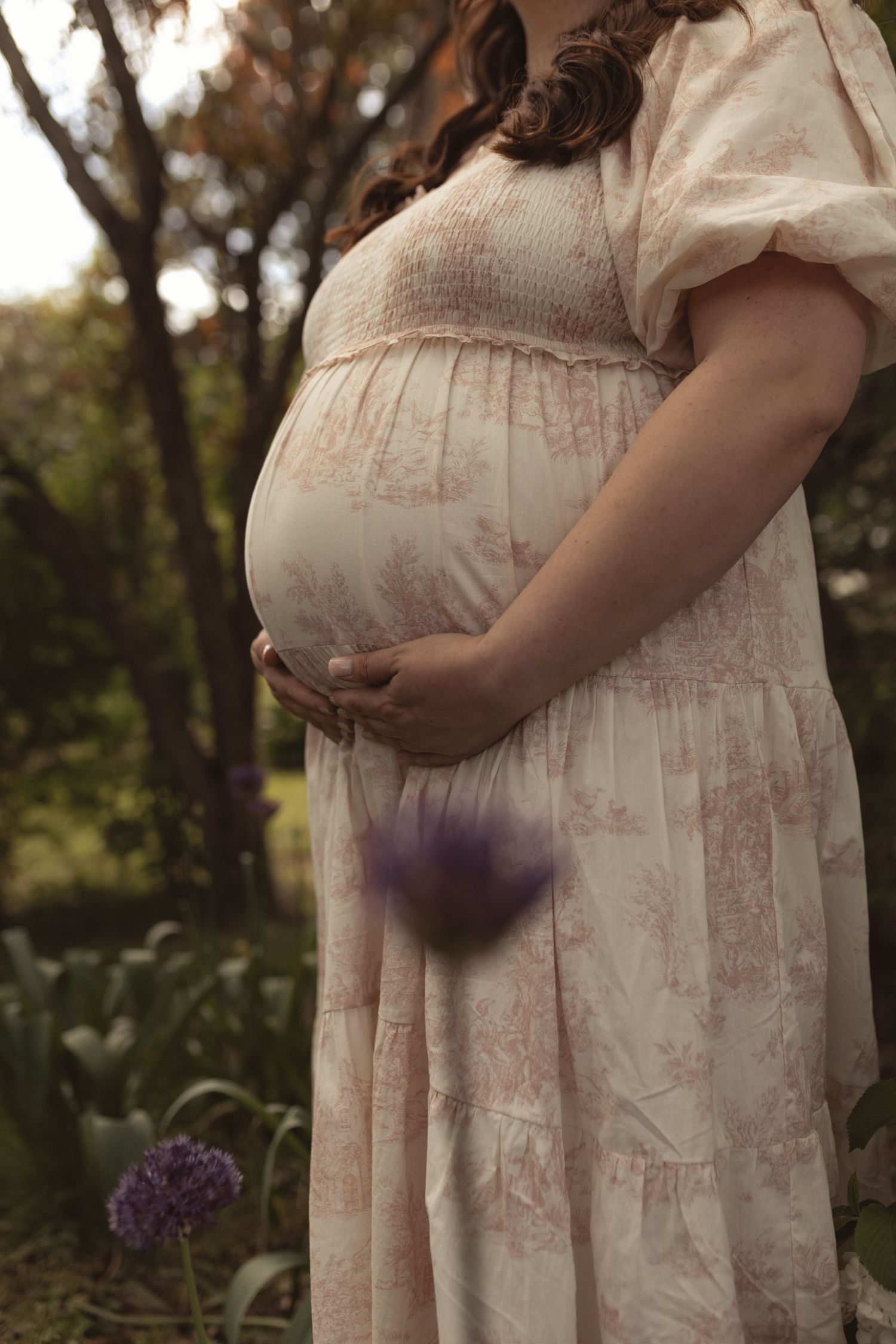 Pregnant woman in floral dress cradling baby bump outdoors with purple allium flowers in a garden setting.