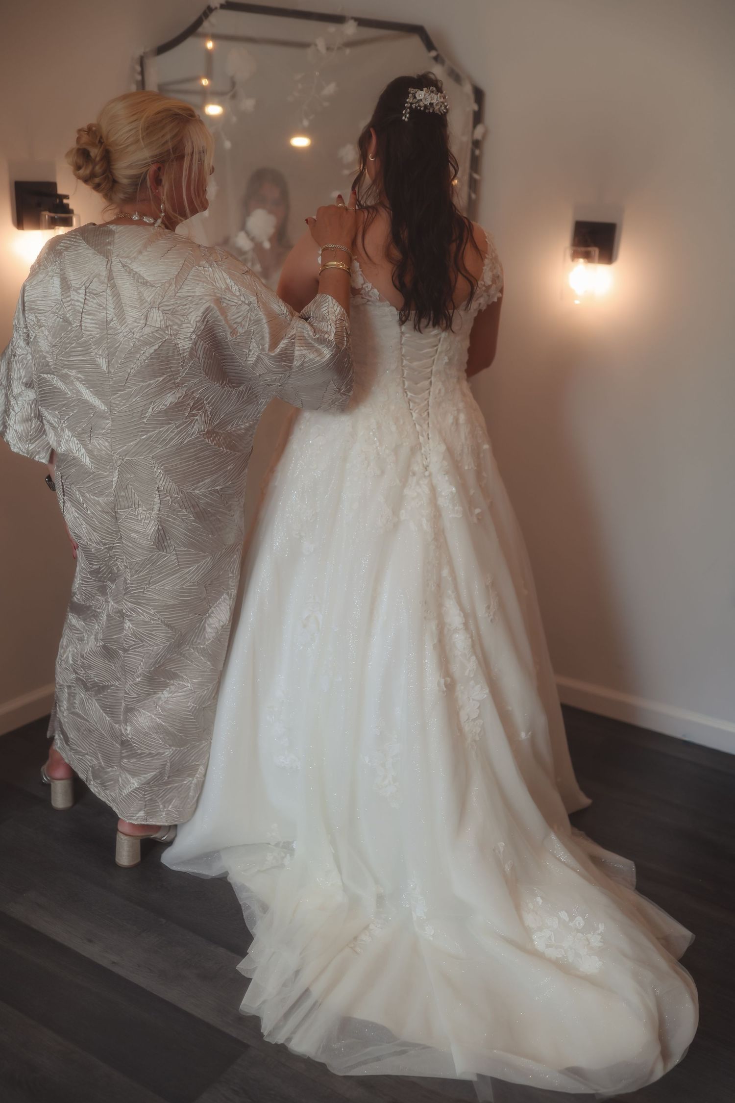 Bride in lace wedding gown with train stands before mirror as woman in silver dress fastens the back.
