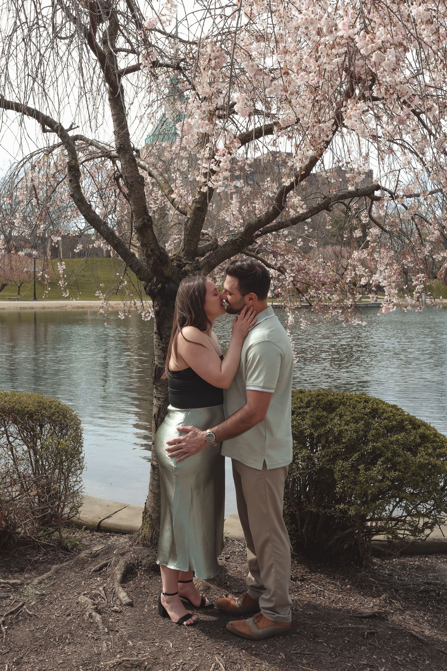 Couple kissing under blooming cherry blossom tree by a serene lake, woman in sage green skirt and black top.