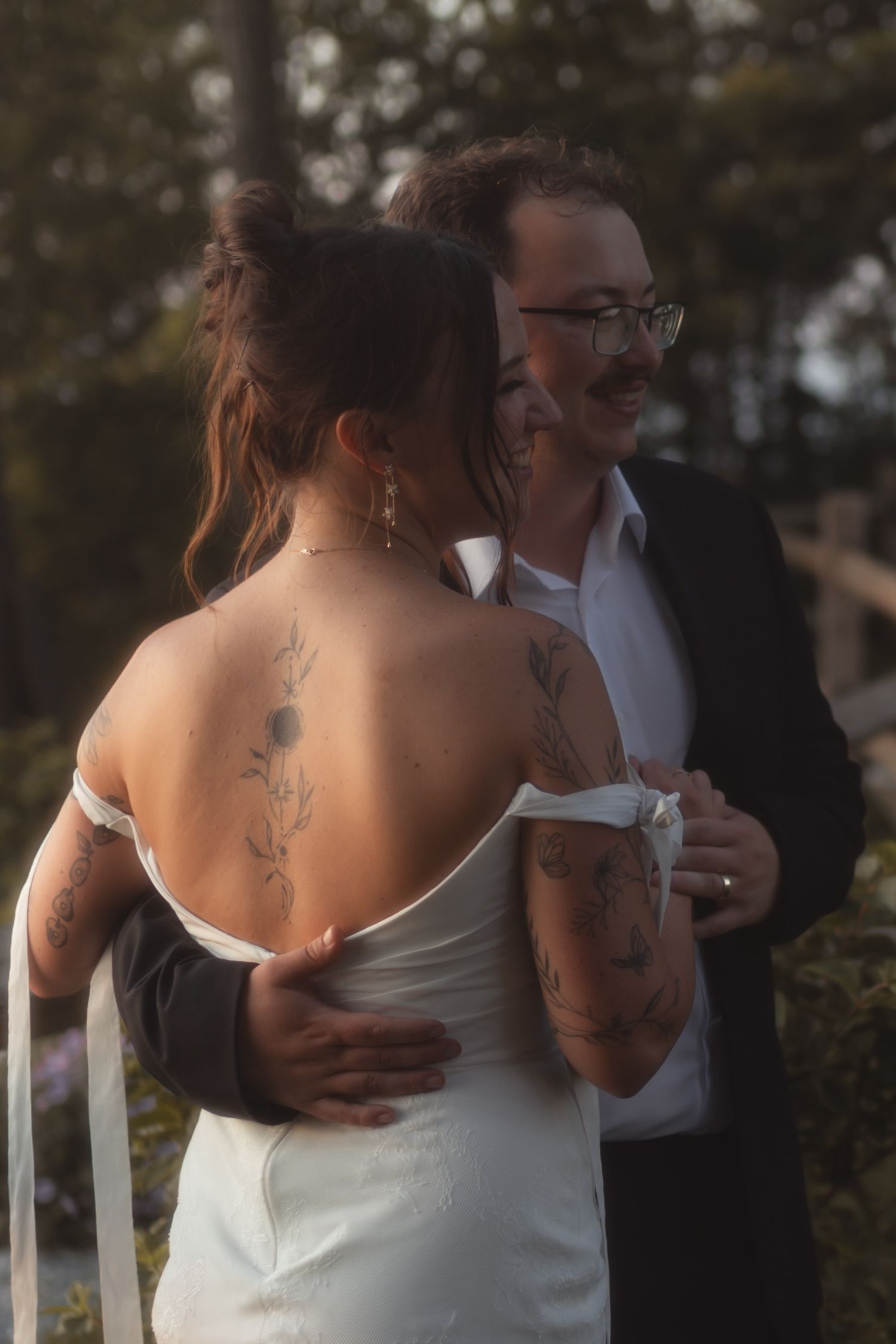 Tattooed bride in backless white gown embraced by groom in black suit during outdoor wedding ceremony.