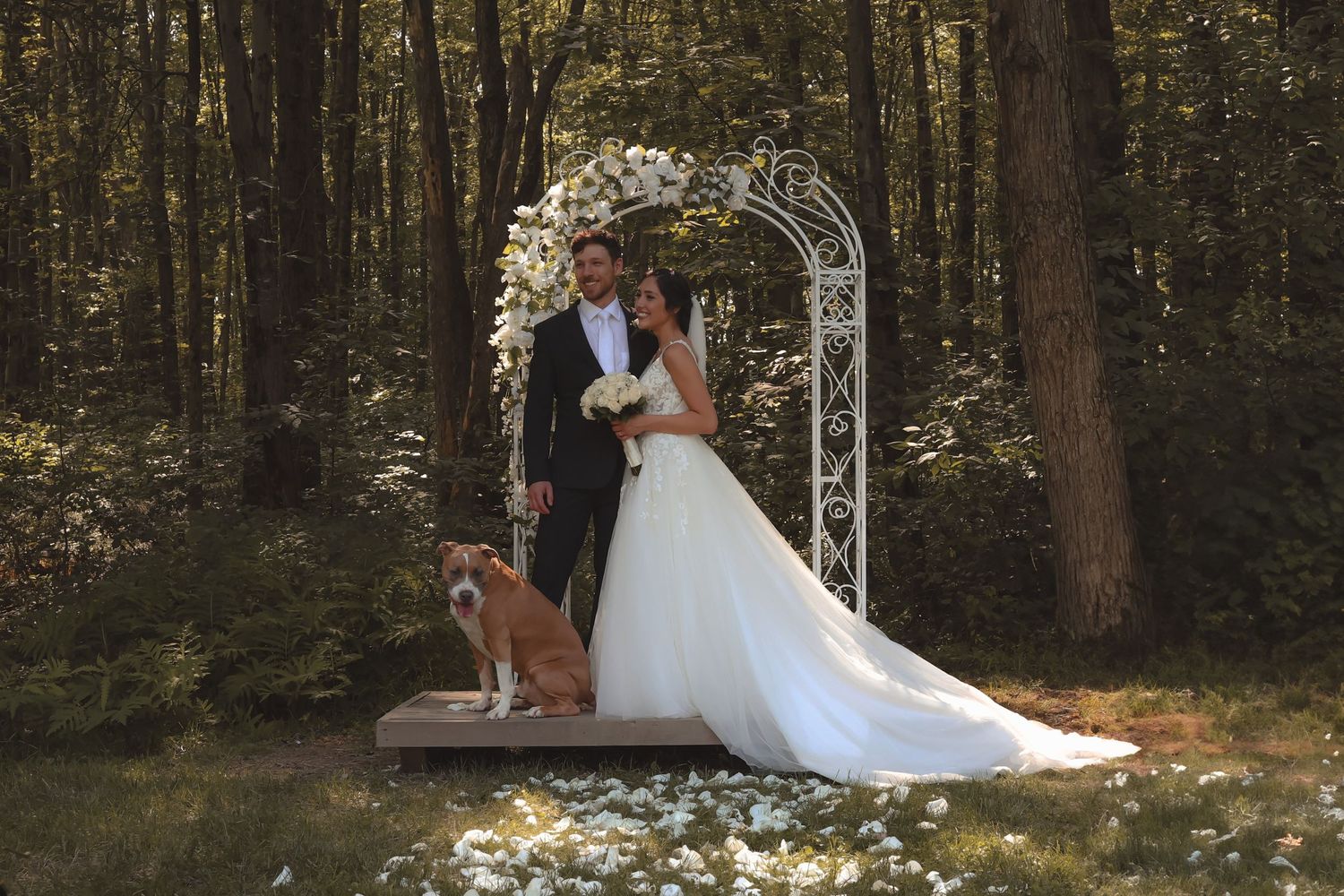 Bride and groom pose under floral arch in forest wedding with their dog beside them.
