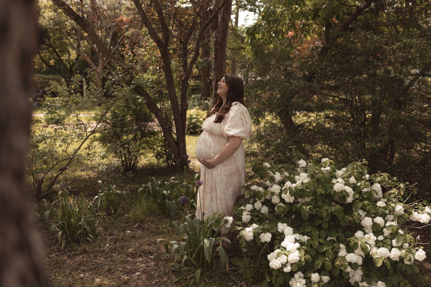 Pregnant woman in white lace dress standing among white flowers in a lush green forest during a maternity photo session.