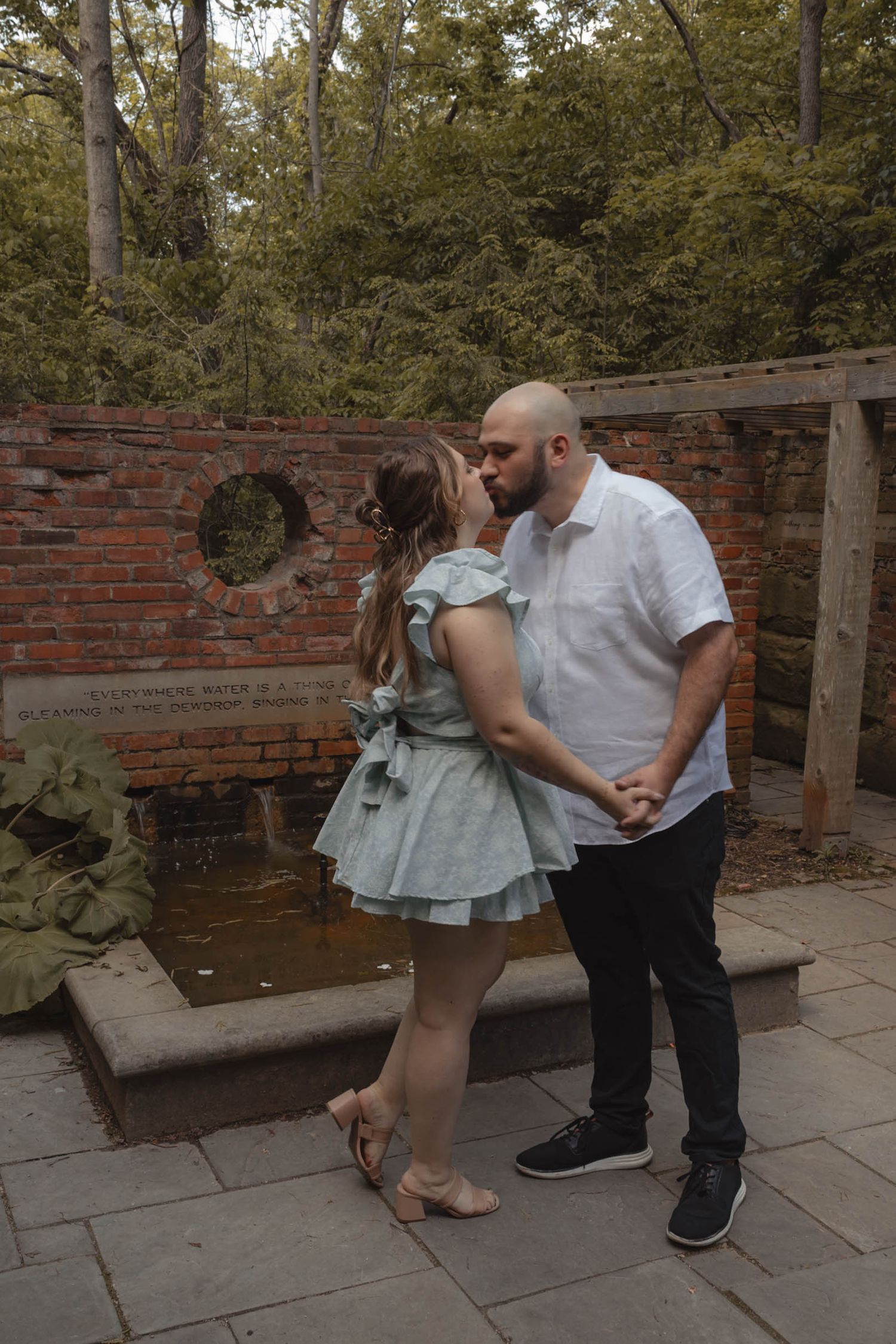 Couple sharing a romantic kiss in a garden courtyard with brick wall and pond backdrop.