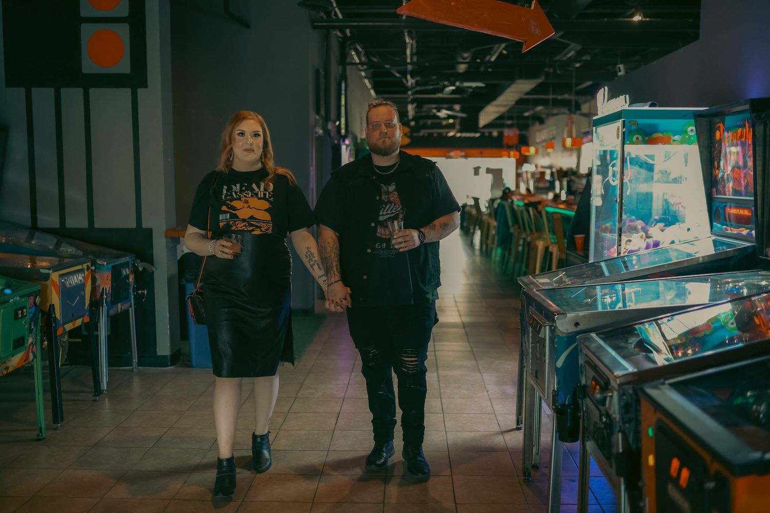 Couple walking hand-in-hand through a retro arcade with colorful pinball machines lining the walls.