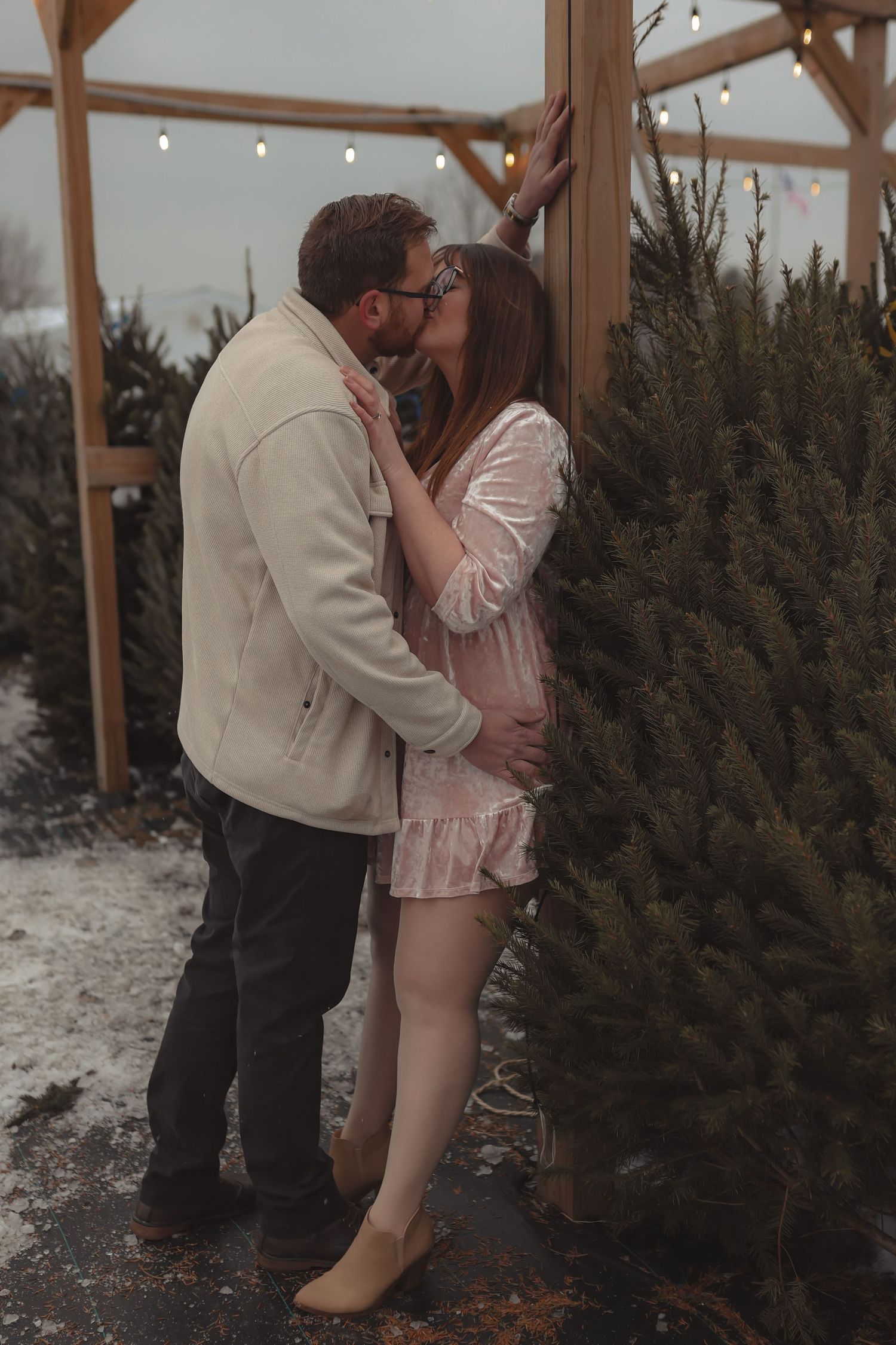 Couple sharing a romantic kiss at a snowy Christmas tree farm, woman in pink dress showing baby bump.