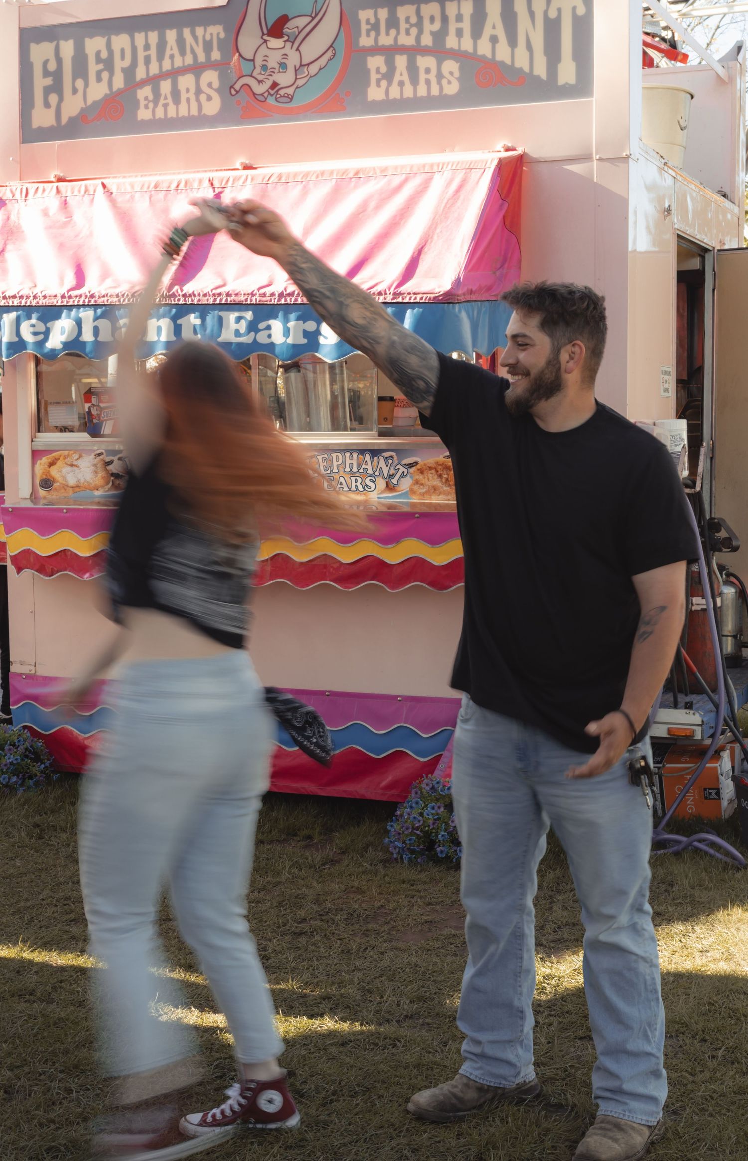 A tattooed couple dances joyfully in front of an Elephant Ears food stand at a carnival.