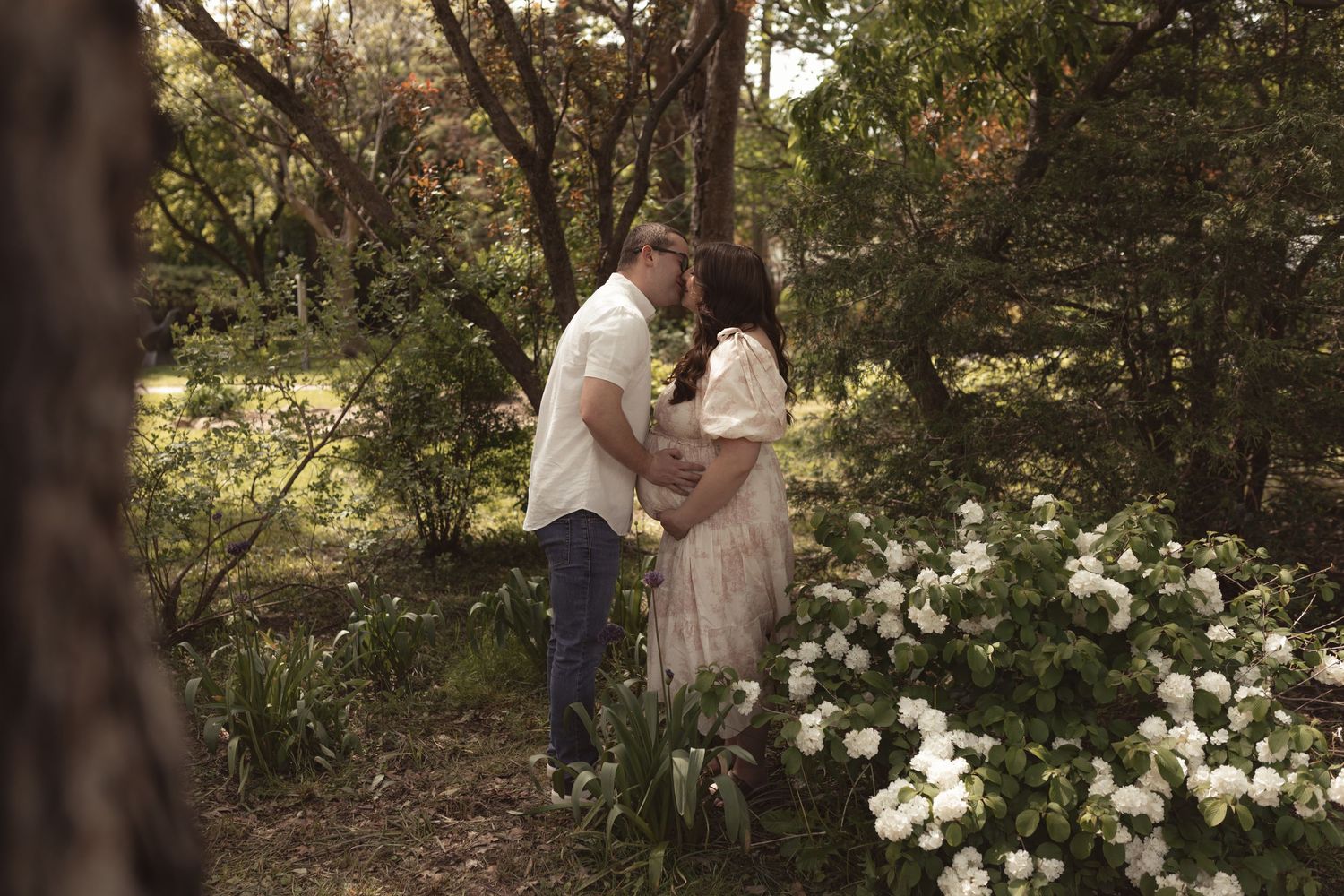 Couple sharing a romantic moment surrounded by white flowers and lush greenery in a serene garden setting.