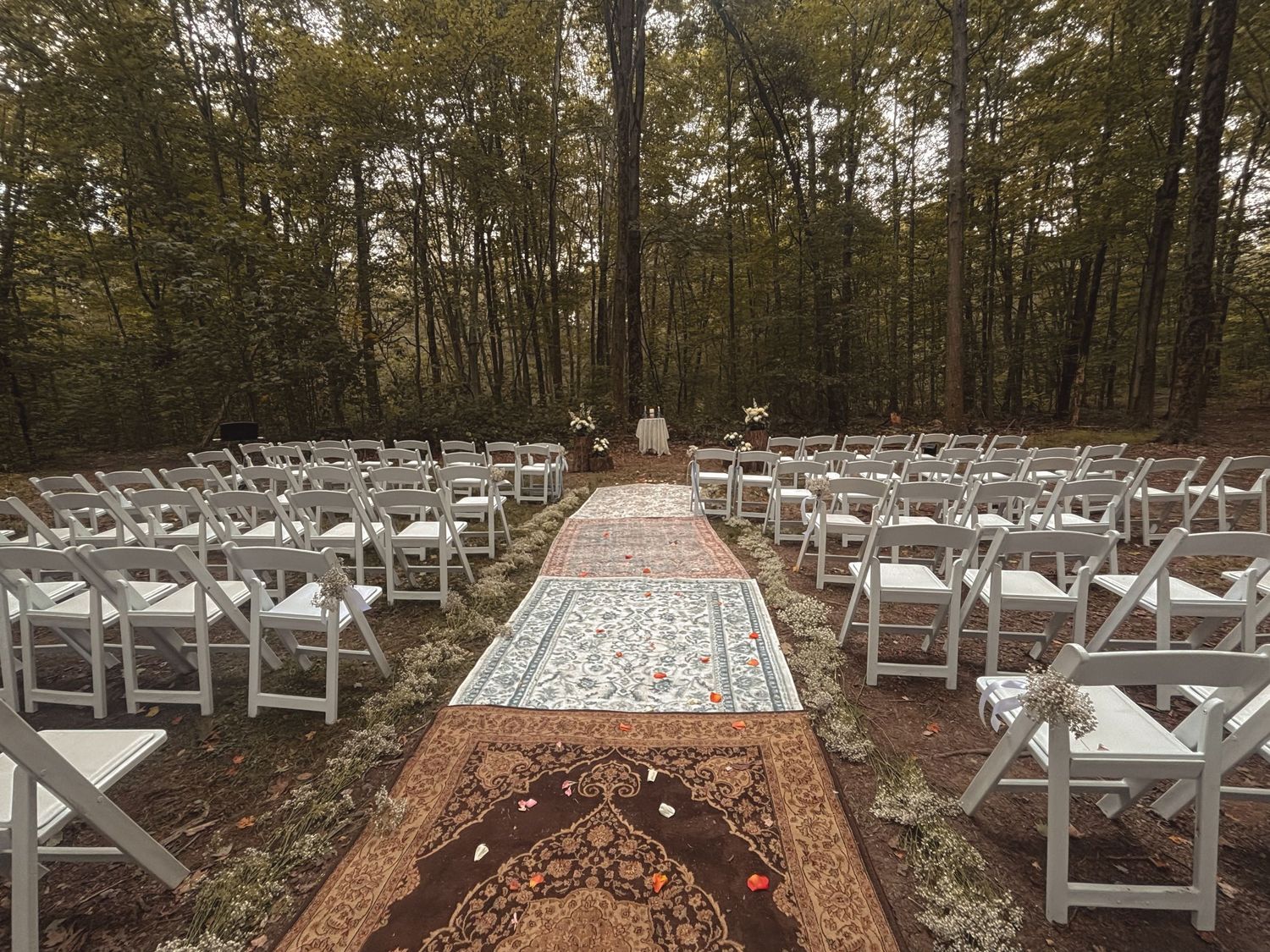 Elegant outdoor woodland wedding ceremony aisle with ornate rugs and rows of white folding chairs.