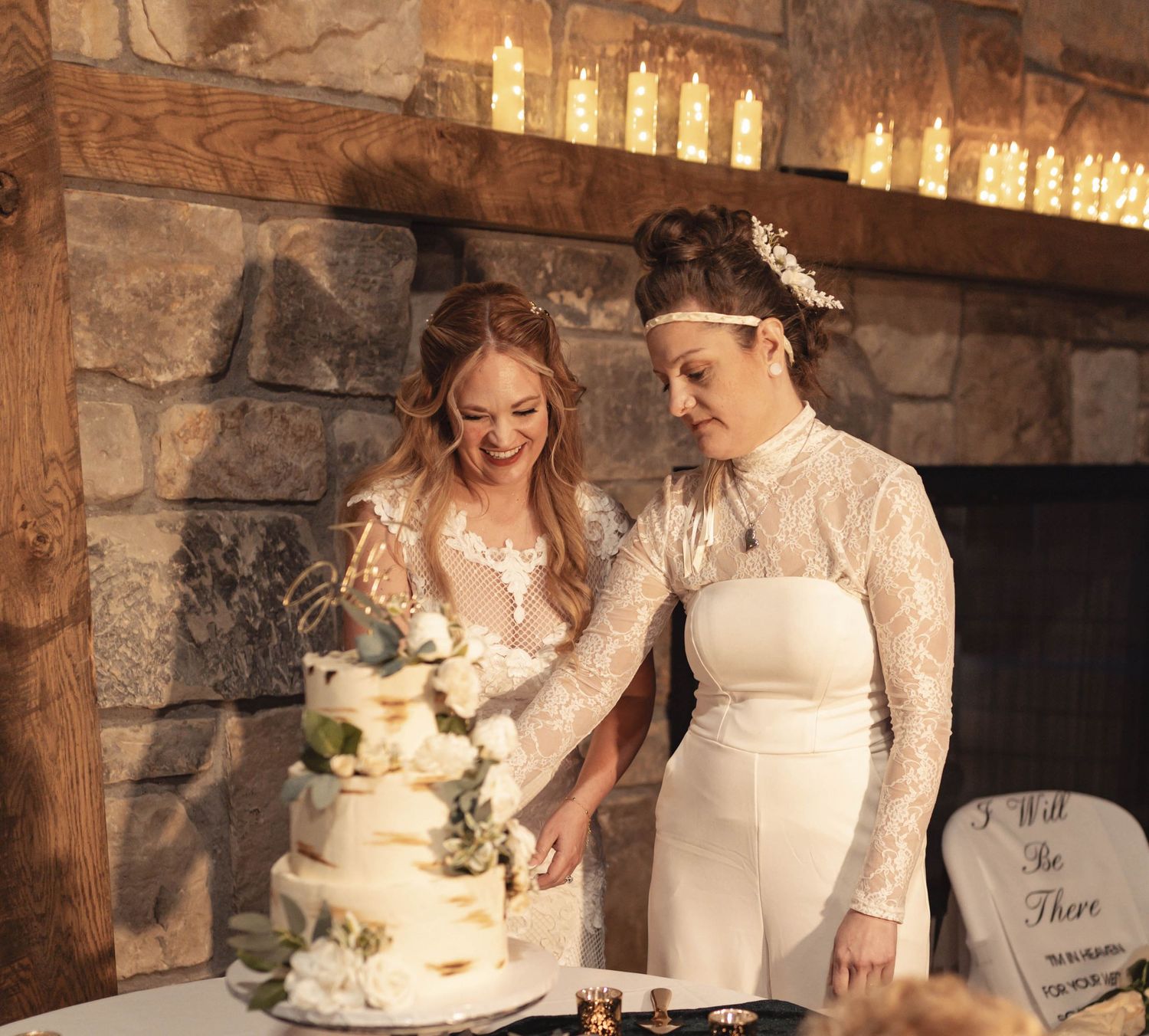 Two brides cutting their elegant white wedding cake together in front of a stone fireplace.