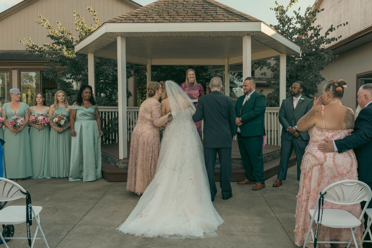 Bride walks down the aisle at an outdoor wedding ceremony near a white gazebo with bridesmaids in sage green.